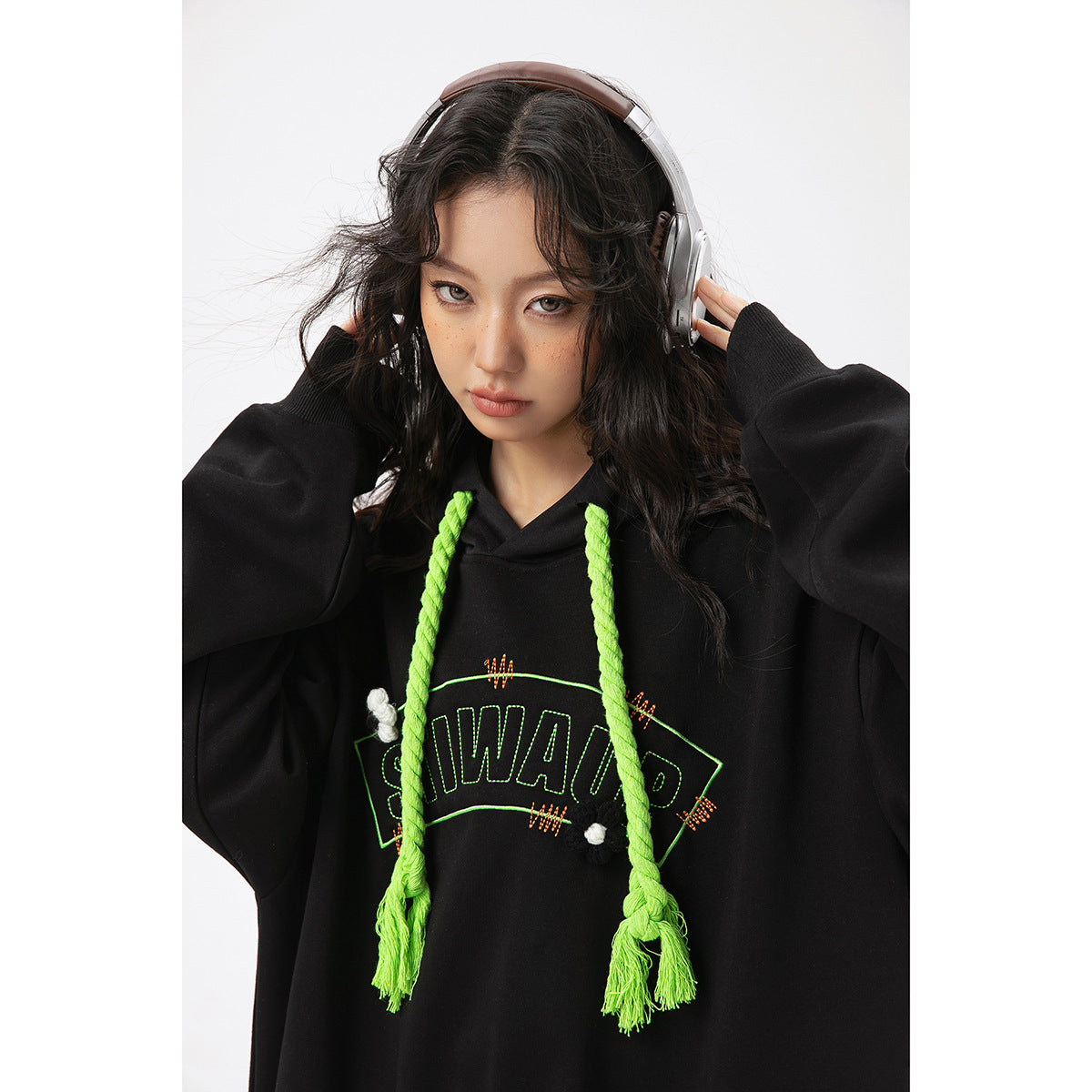 Close view of a model in a black oversized hoodie with neon green braided drawstrings and outlined chest lettering.