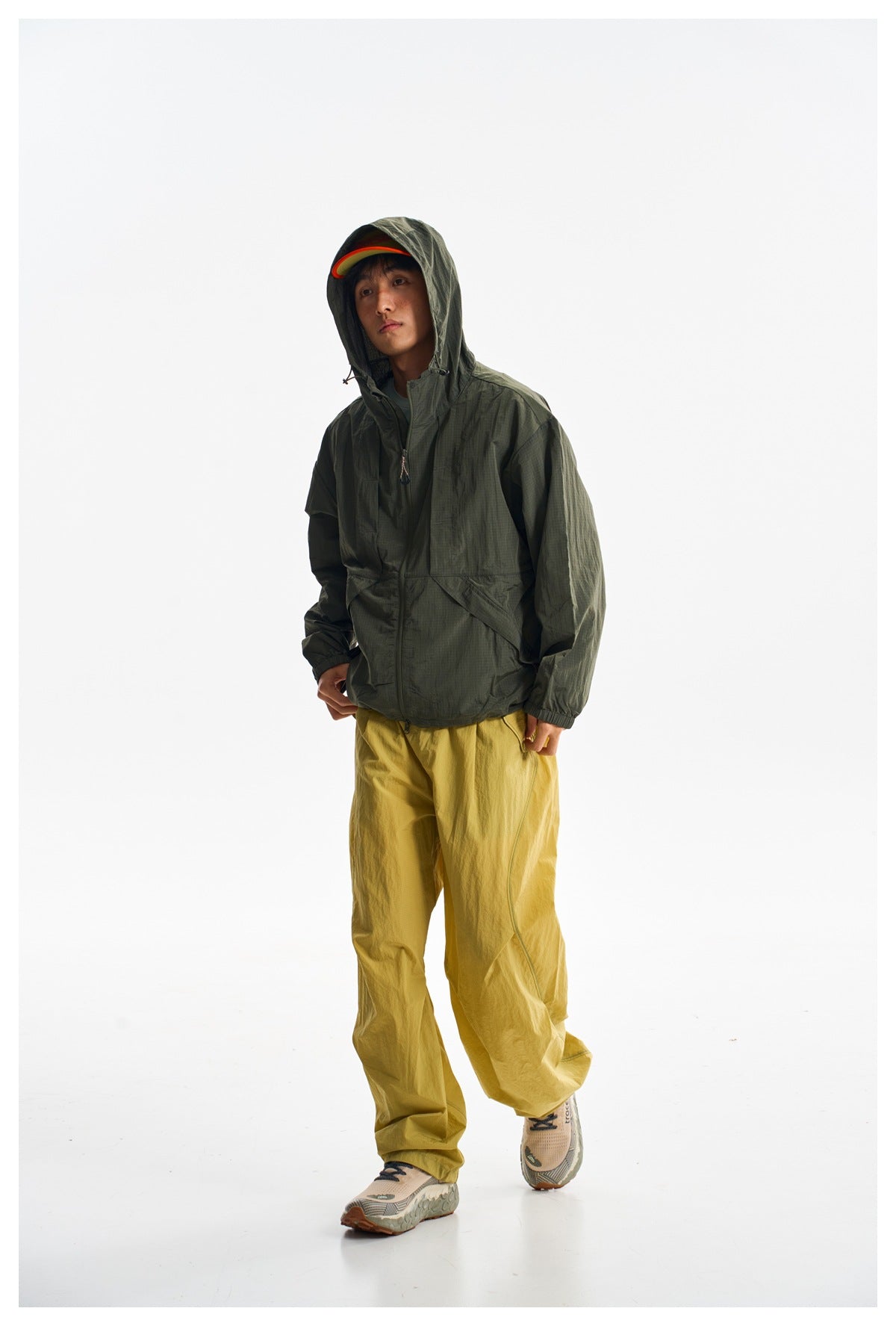 Model wearing a dark olive jacket with the hood up and loose mustard-yellow trousers against a white studio background.