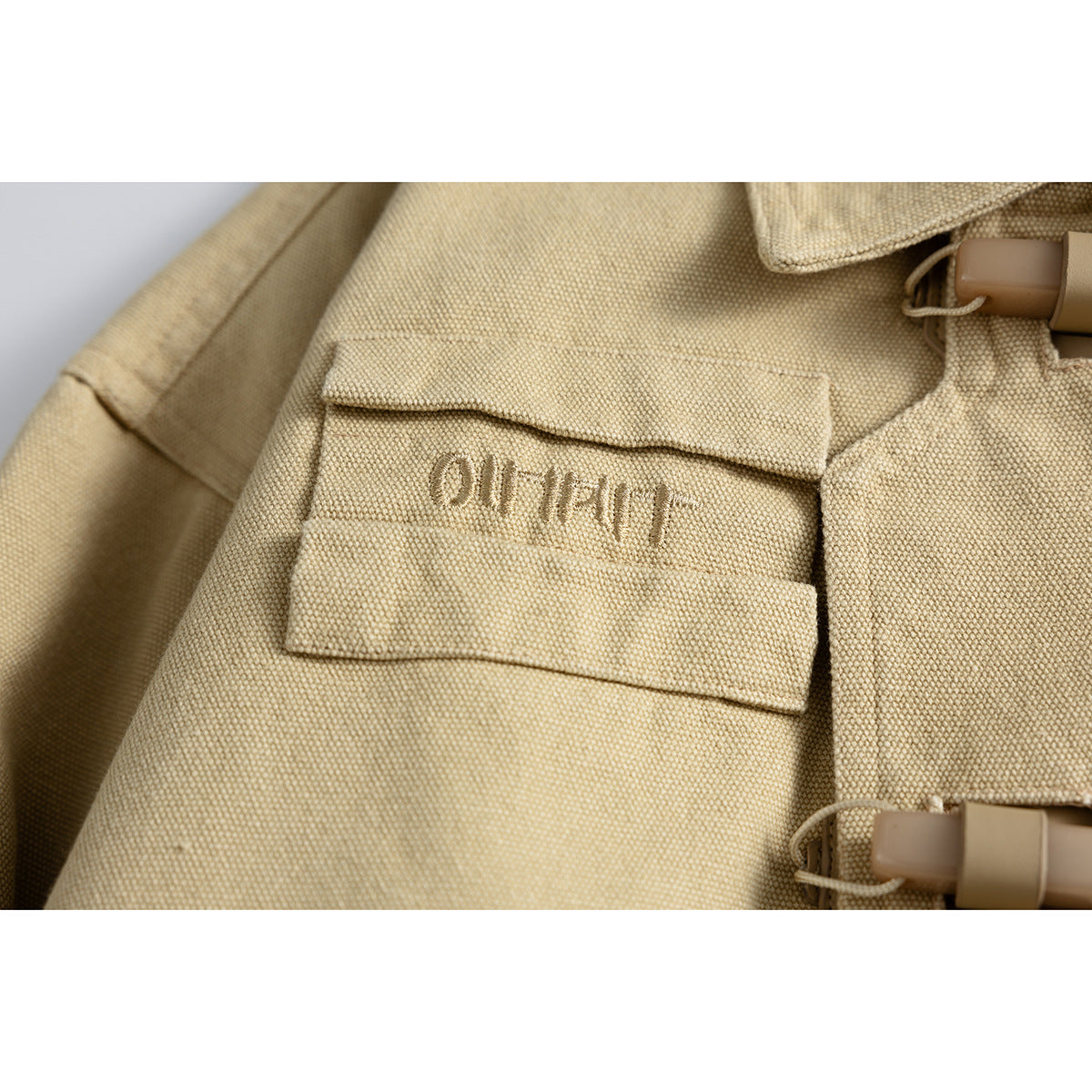 Close-up of a sand-colored jacket's flap chest pocket with subtle embroidery and toggle closures.