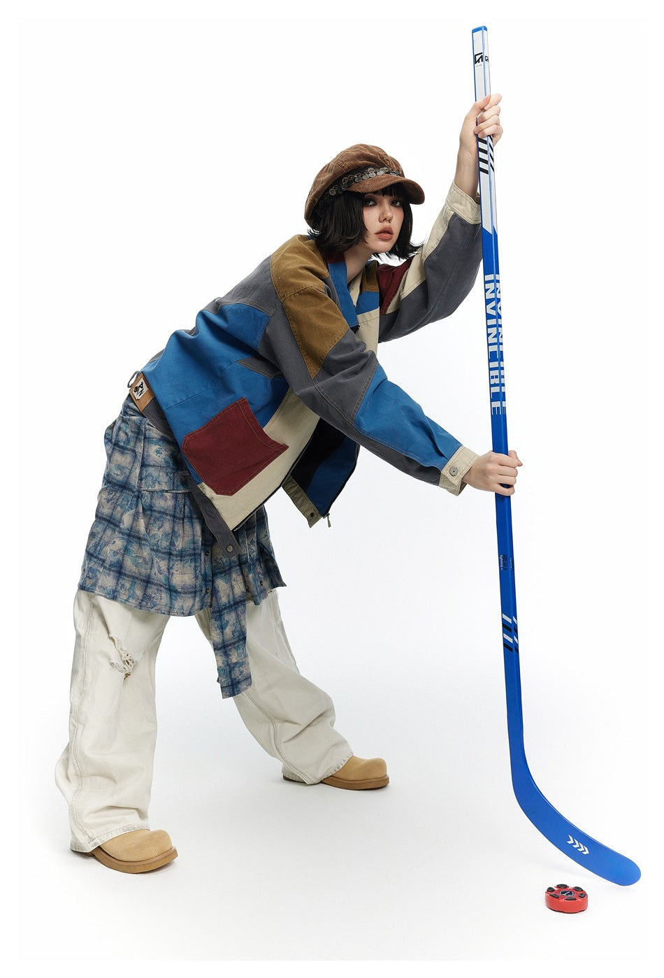 Model wearing a multicolor patchwork jacket with a plaid layer and cream trousers, holding a blue hockey stick.