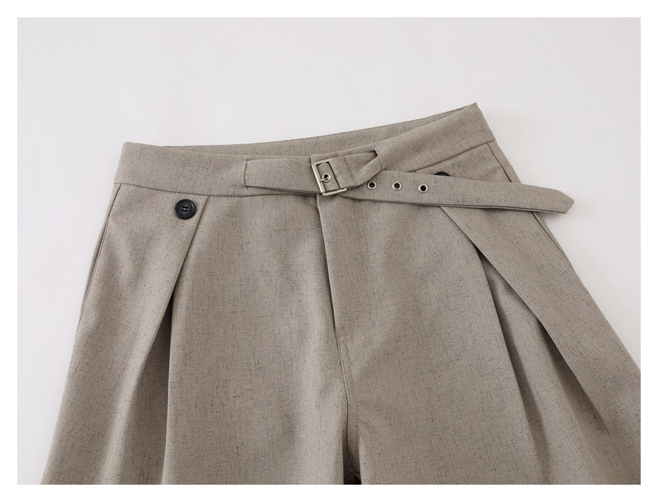Close-up of taupe trousers showing the front waistband, button, pleats, and adjustable buckle belt.