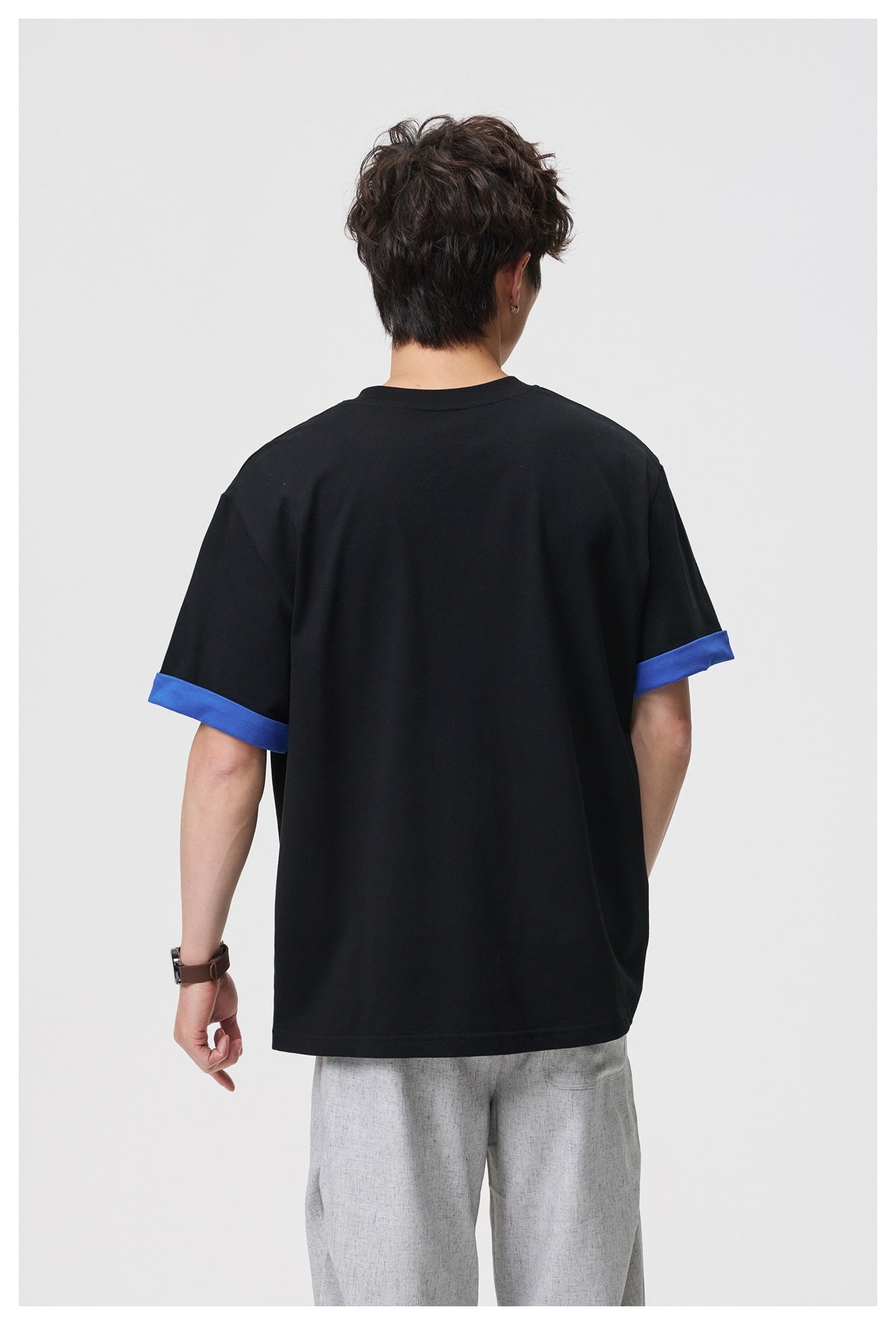 Back view of a model wearing a black T-shirt with blue turned-up sleeve cuffs and light gray trousers.