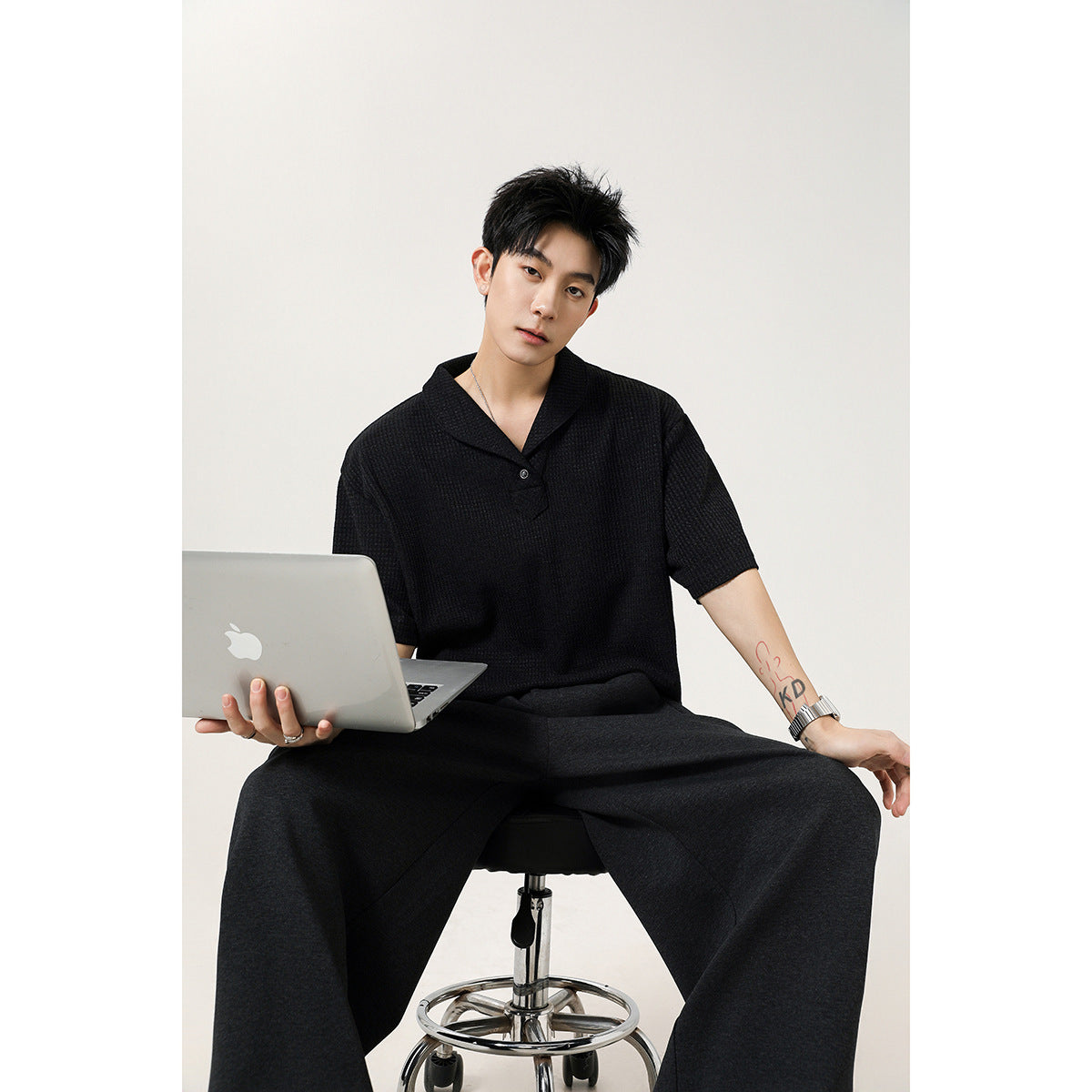 Seated model wearing a black short-sleeve knit polo shirt with a shawl collar and dark trousers.