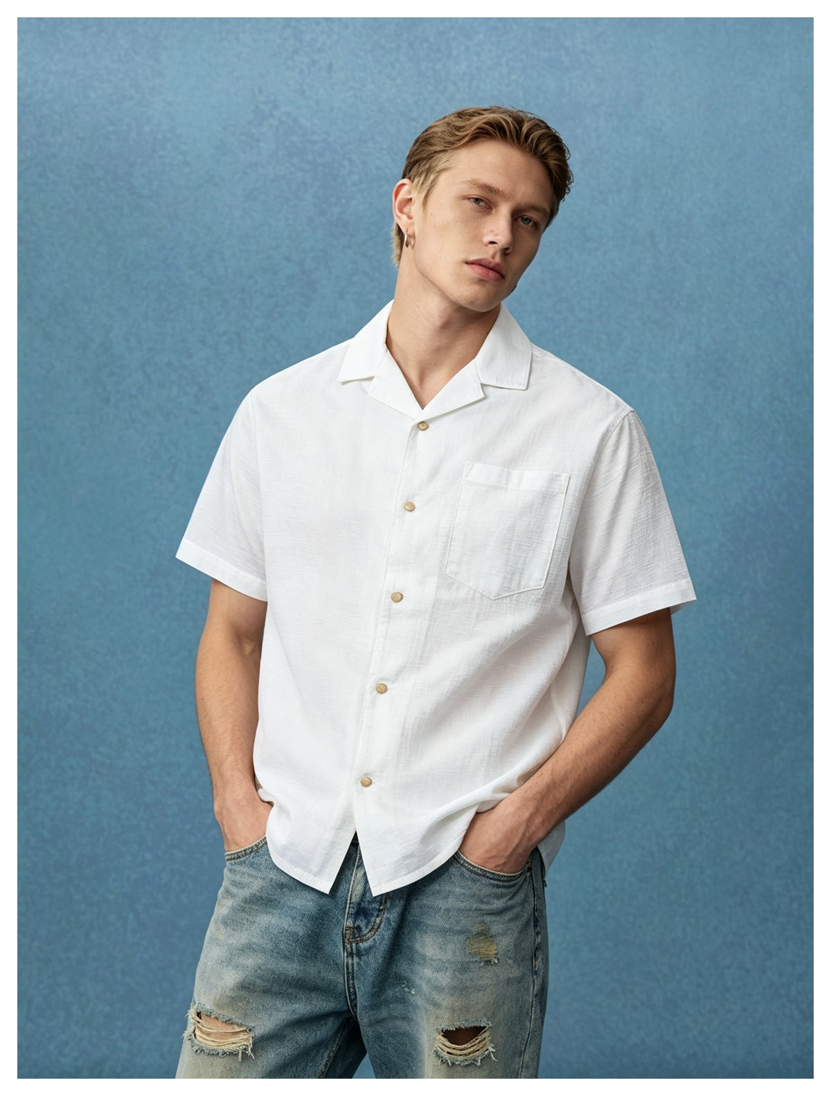 Man wearing a white short-sleeve camp collar shirt with chest pocket and ripped jeans against a blue backdrop