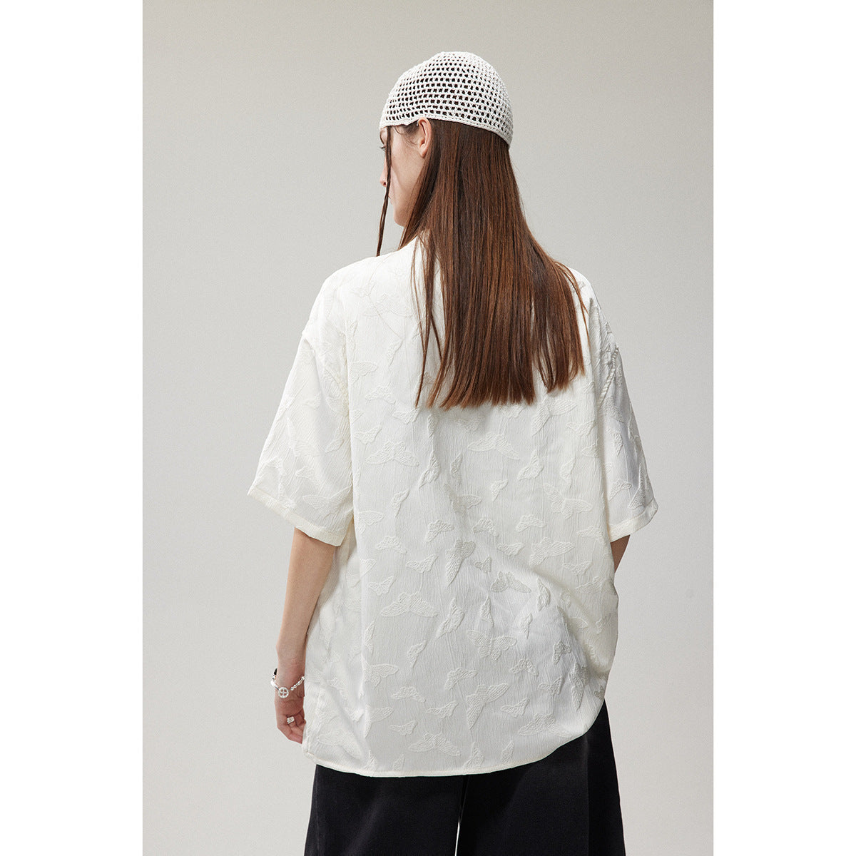 Back view of a model wearing a white short-sleeve shirt with raised butterfly motifs and black trousers.