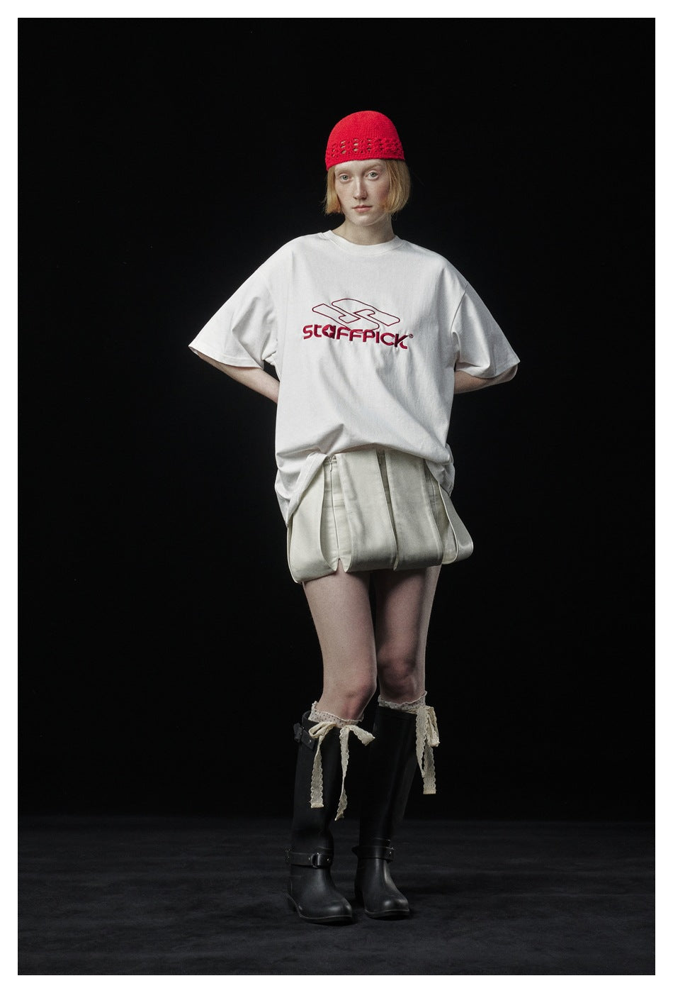 Front view of a model wearing a white graphic T-shirt, pale pleated shorts, a red knit cap, and black boots.