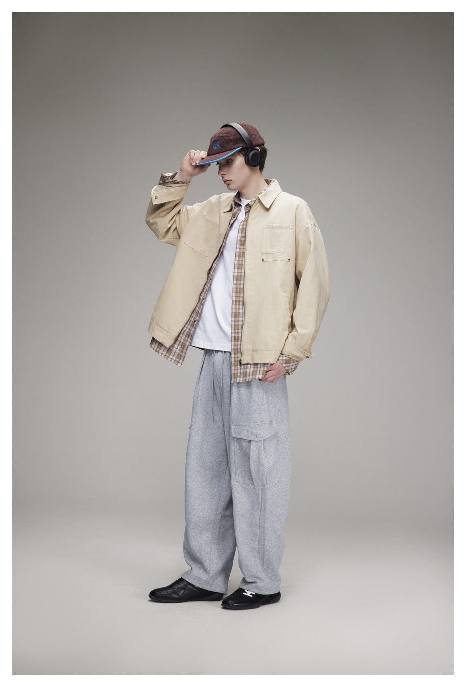 Model in an open khaki zip jacket over a plaid shirt and white tee with gray cargo pants, hand on cap