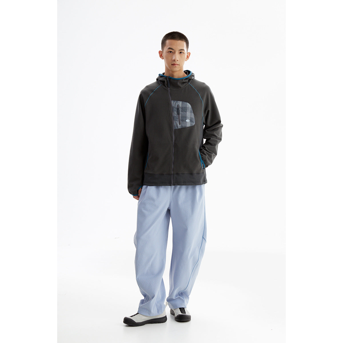 Model wearing a charcoal zip-up hoodie with blue piping and a plaid chest pocket, paired with loose light-blue pants.