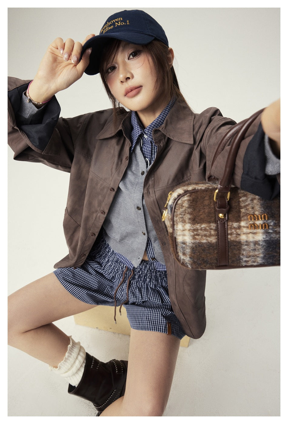 Model in a brown button-front shirt jacket over a gray vest, plaid shirt and shorts, holding a plaid handbag