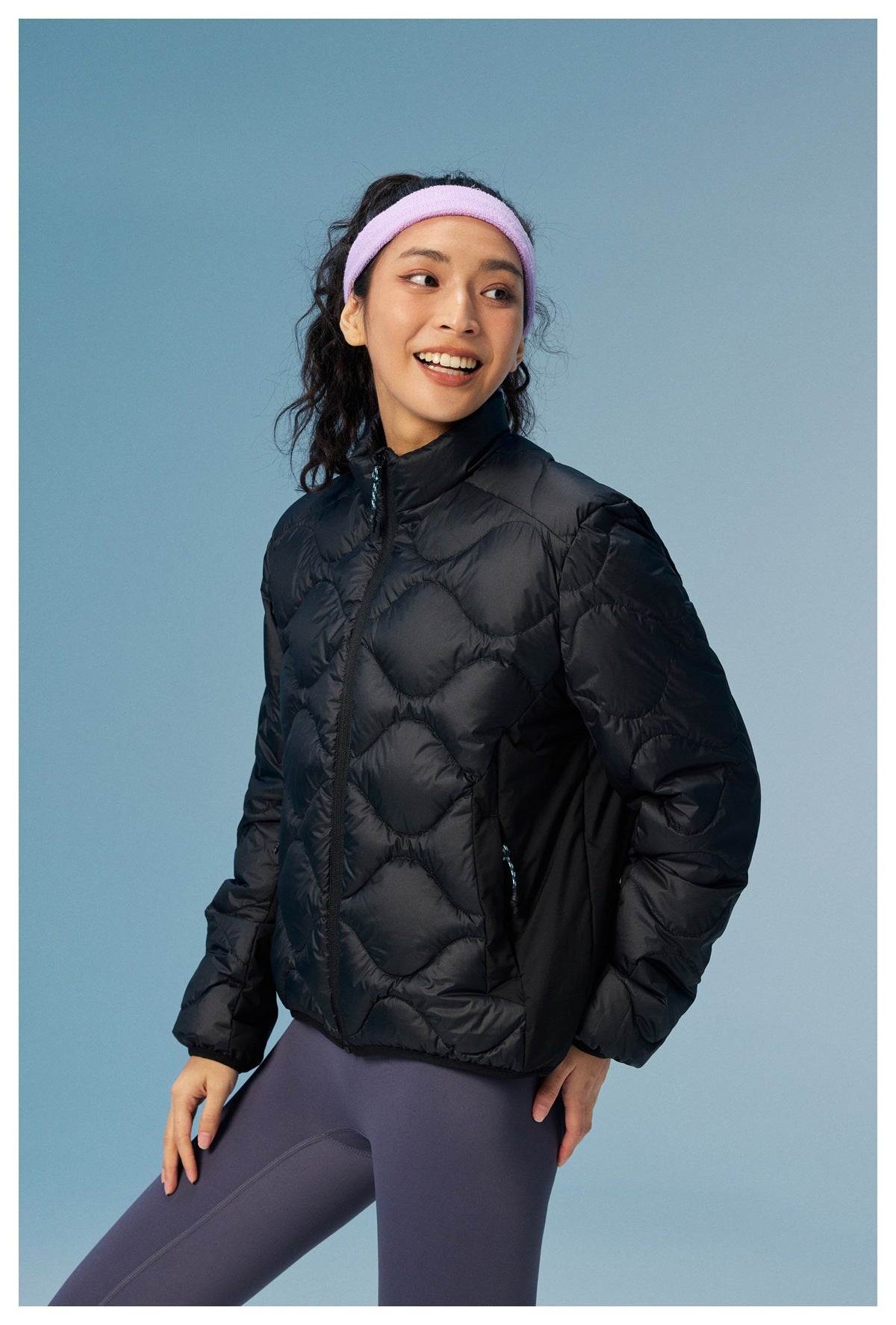 Three-quarter view of a woman wearing a black wave-quilted zip jacket and purple leggings.
