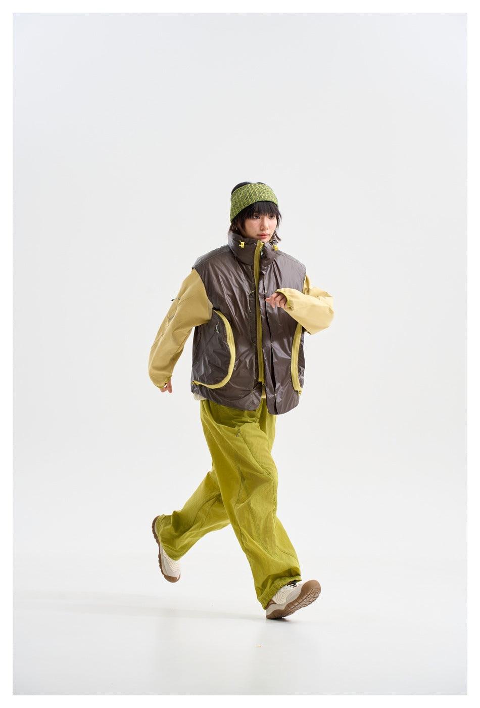 Model walking in a glossy charcoal vest with yellow-trimmed pockets, a pale yellow top and lime-green trousers.