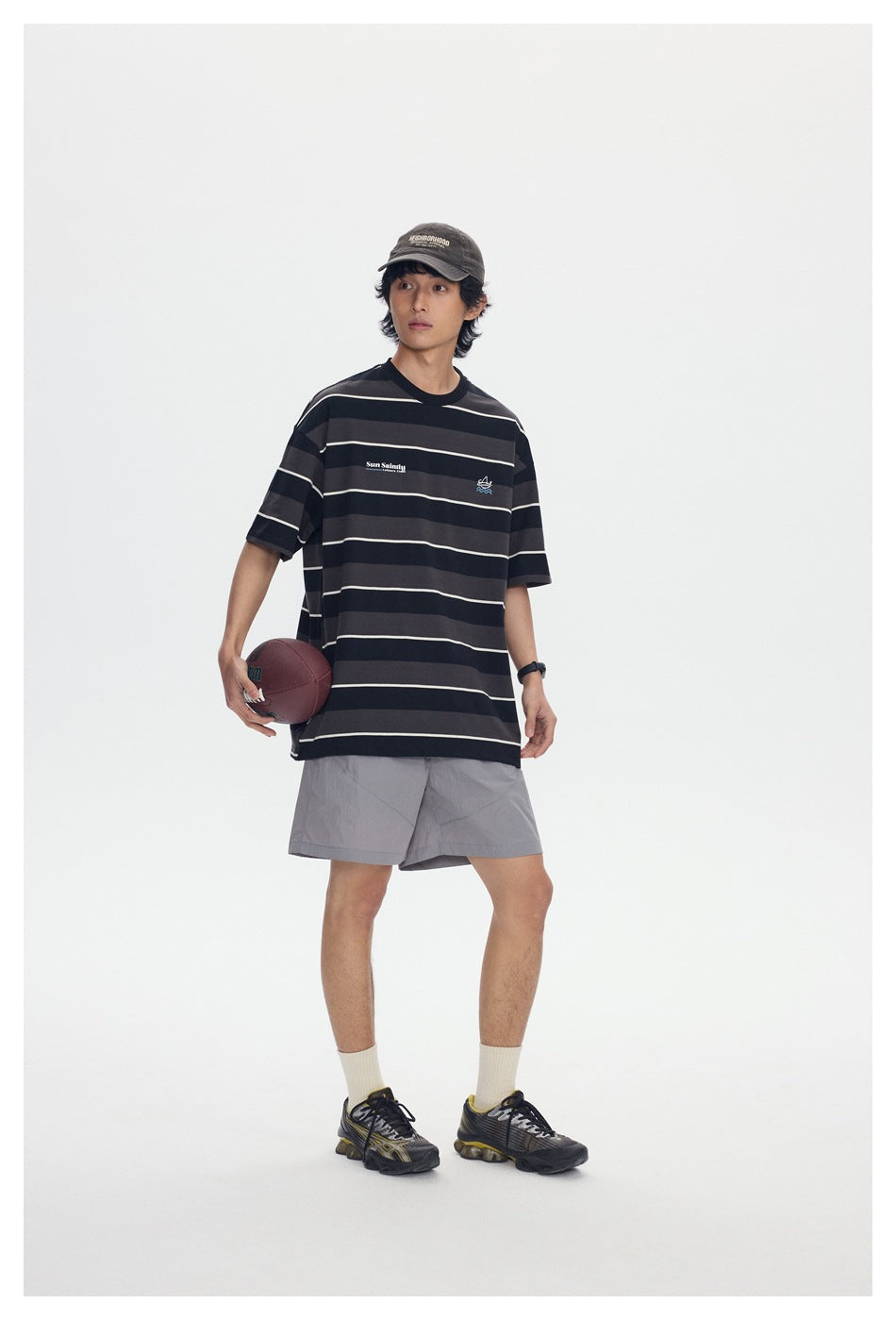 Model wearing a black and charcoal striped T-shirt with gray shorts, cap, and sneakers.