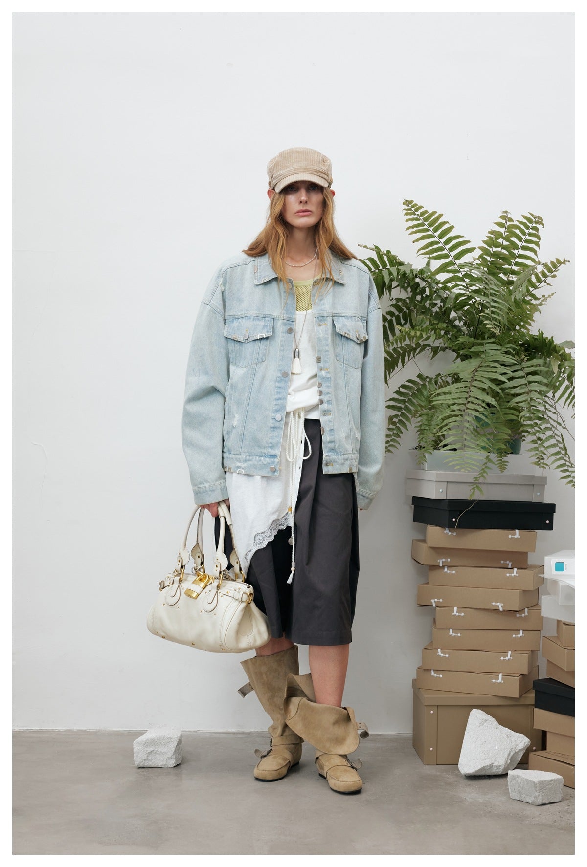 Model wearing an oversized light blue denim jacket over a white layer, dark skirt, cap and suede boots, facing front