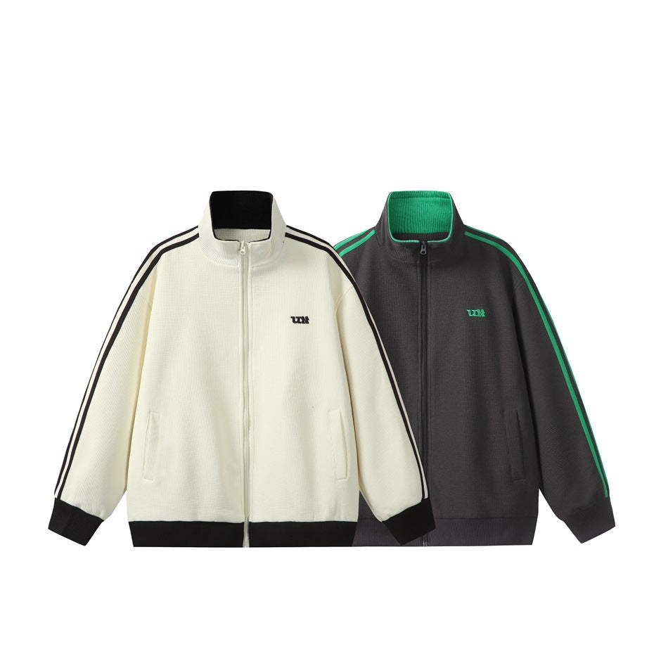 Two zip-up jackets shown from the front: ivory with black stripes and trim, and charcoal with green stripes and trim.