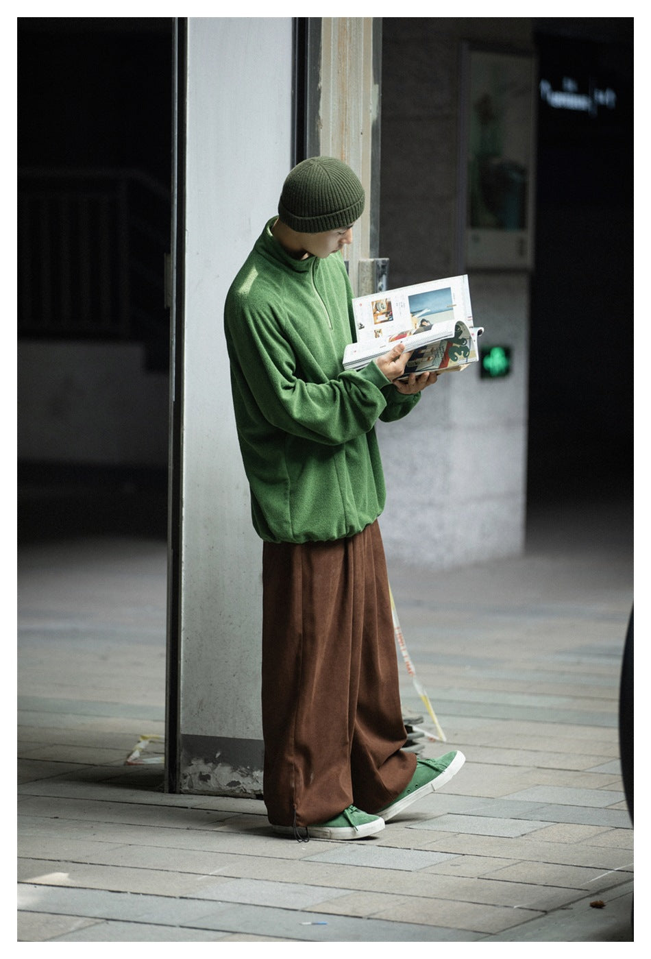 Side view of a model reading a magazine in brown wide-leg pants, a green pullover, a beanie and green sneakers
