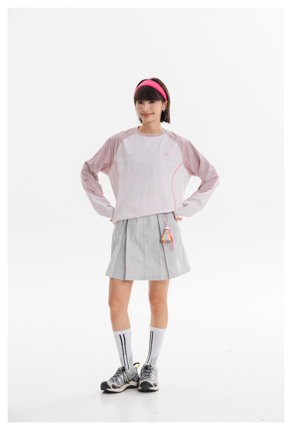 Model wearing a pale pink and white long-sleeve top with a light gray pleated mini skirt, hands on hips.