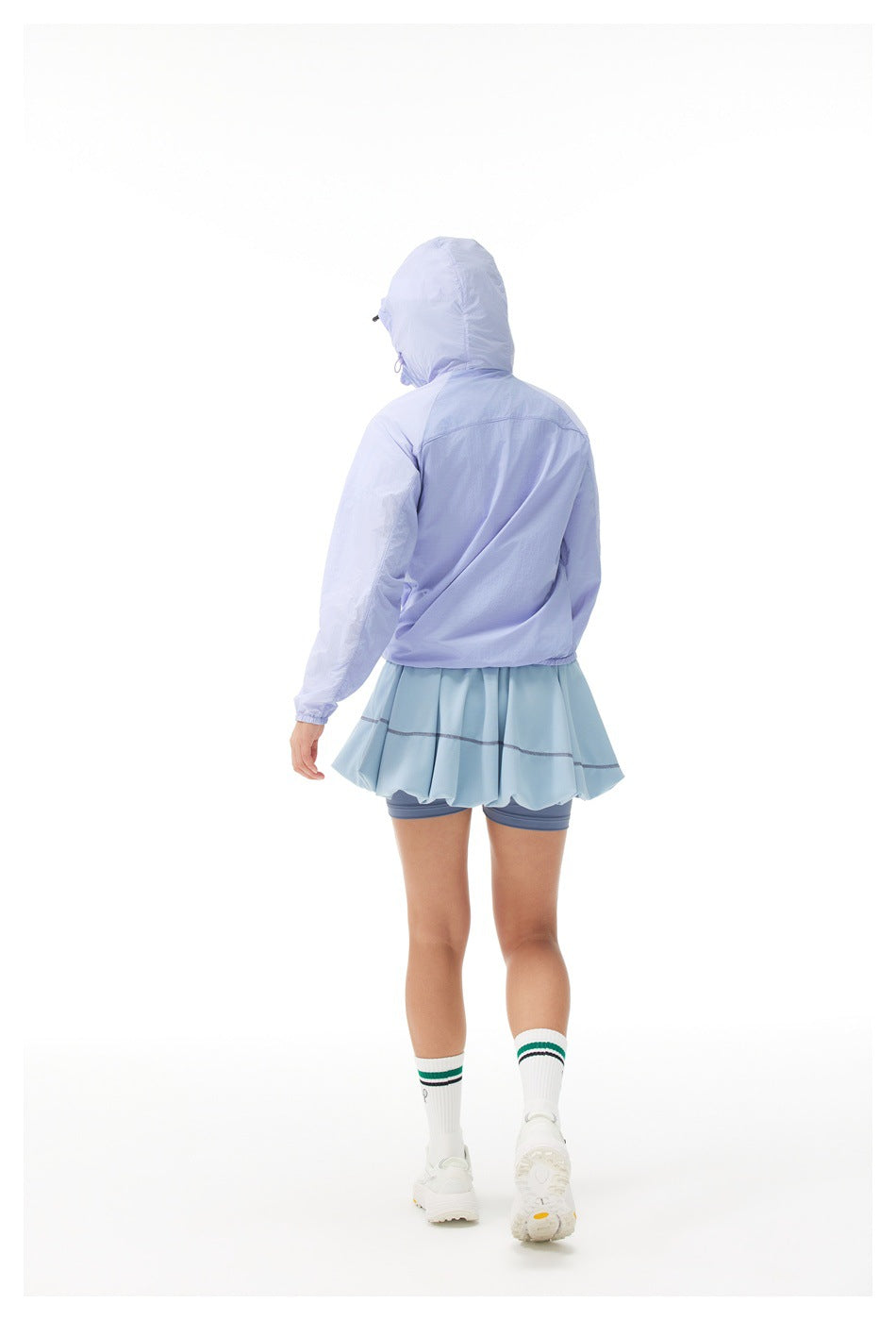 Back view of a woman wearing a purple hooded jacket with the hood up, a light blue skirt and white sneakers