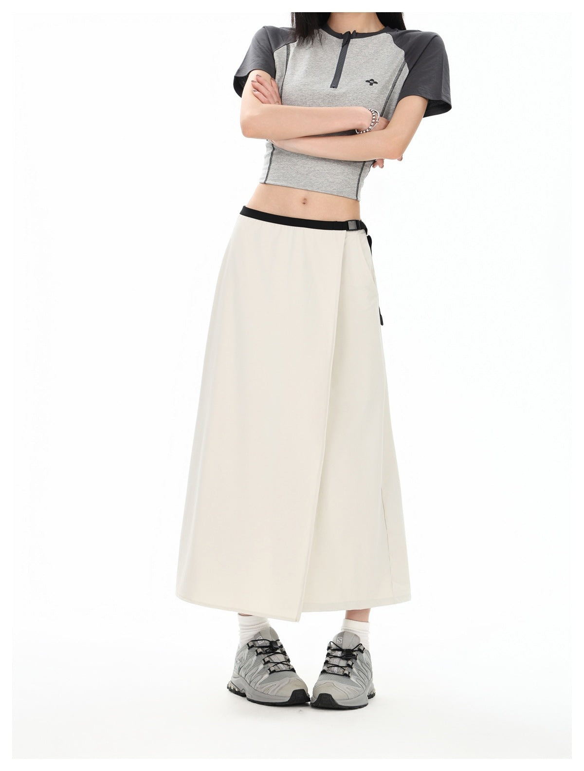 Model wearing a cream wrap-front midi skirt with black waistband and buckle, gray raglan crop top and gray sneakers