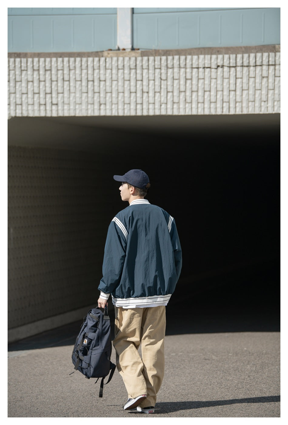 A model walks away wearing a dark teal striped-trim jacket and beige trousers, carrying a dark backpack.