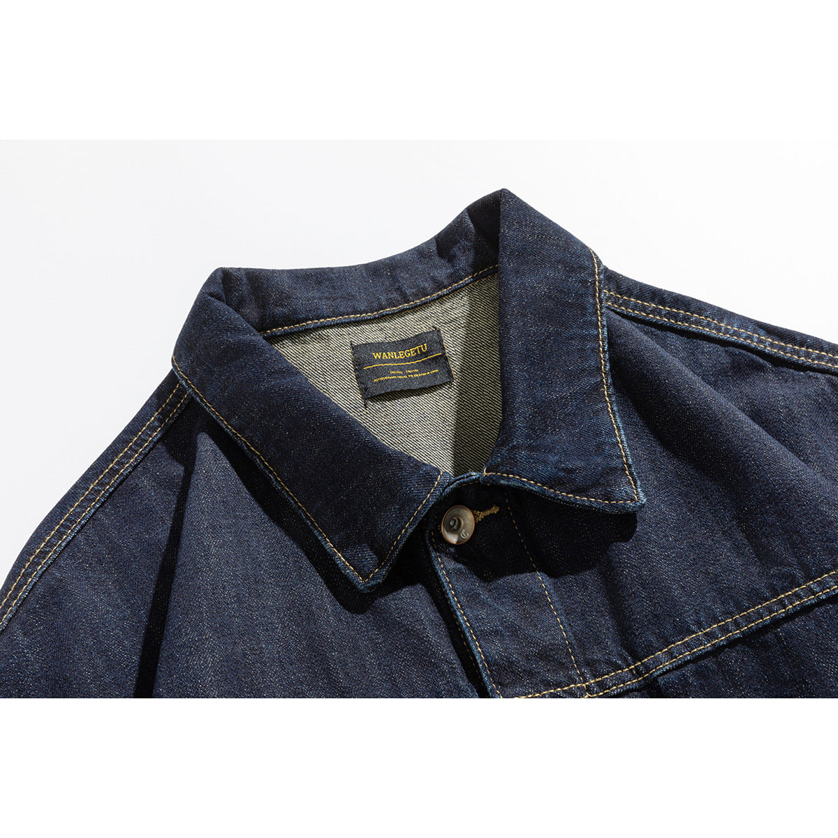 Close-up of a dark indigo denim jacket collar, sewn neck label, button, and golden contrast stitching.