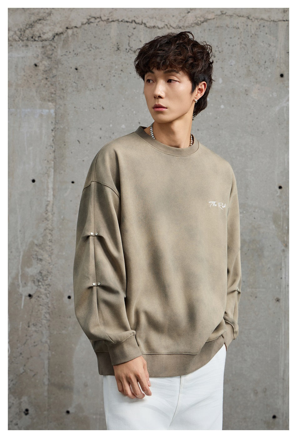 Model wearing a taupe oversized sweatshirt with white chest script and studded sleeve details.