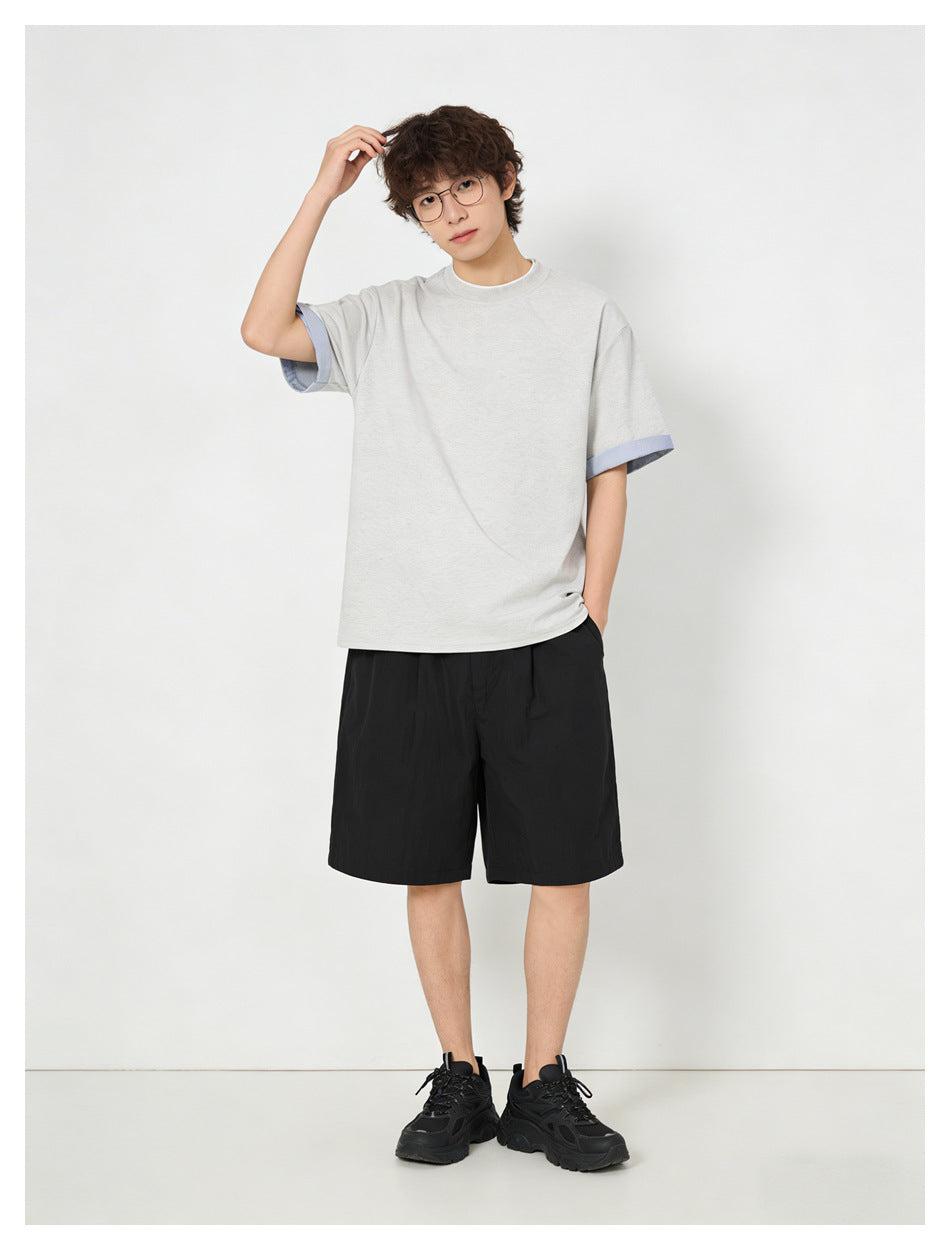 Model wearing a light gray short-sleeve T-shirt with blue cuffs, black shorts, and black sneakers, front view