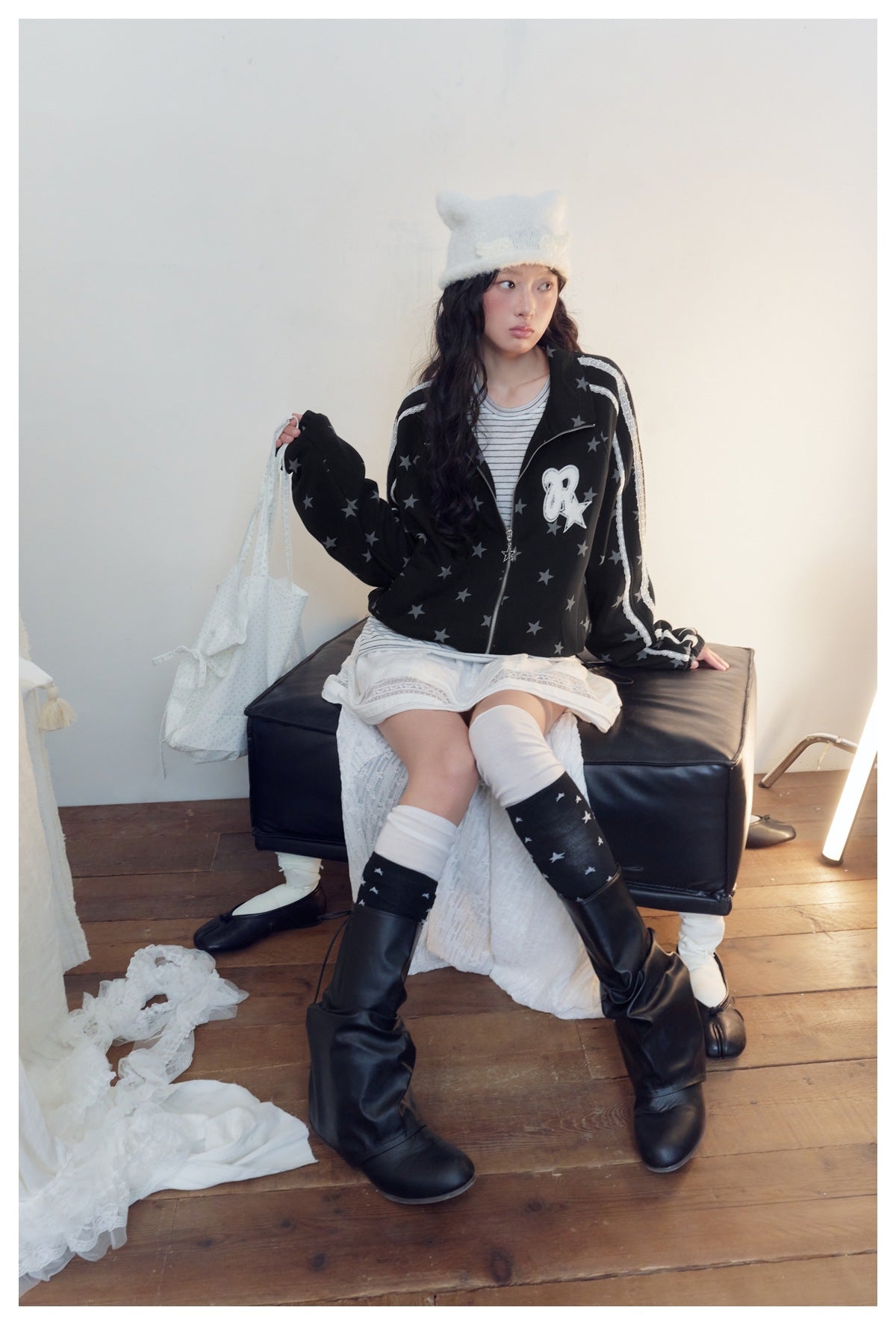 Seated model wearing a black star-print zip jacket with white lace sleeve stripes and a chest patch.