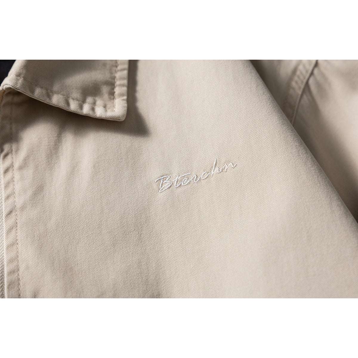 Close-up of white cursive embroidery on the chest of a beige jacket.