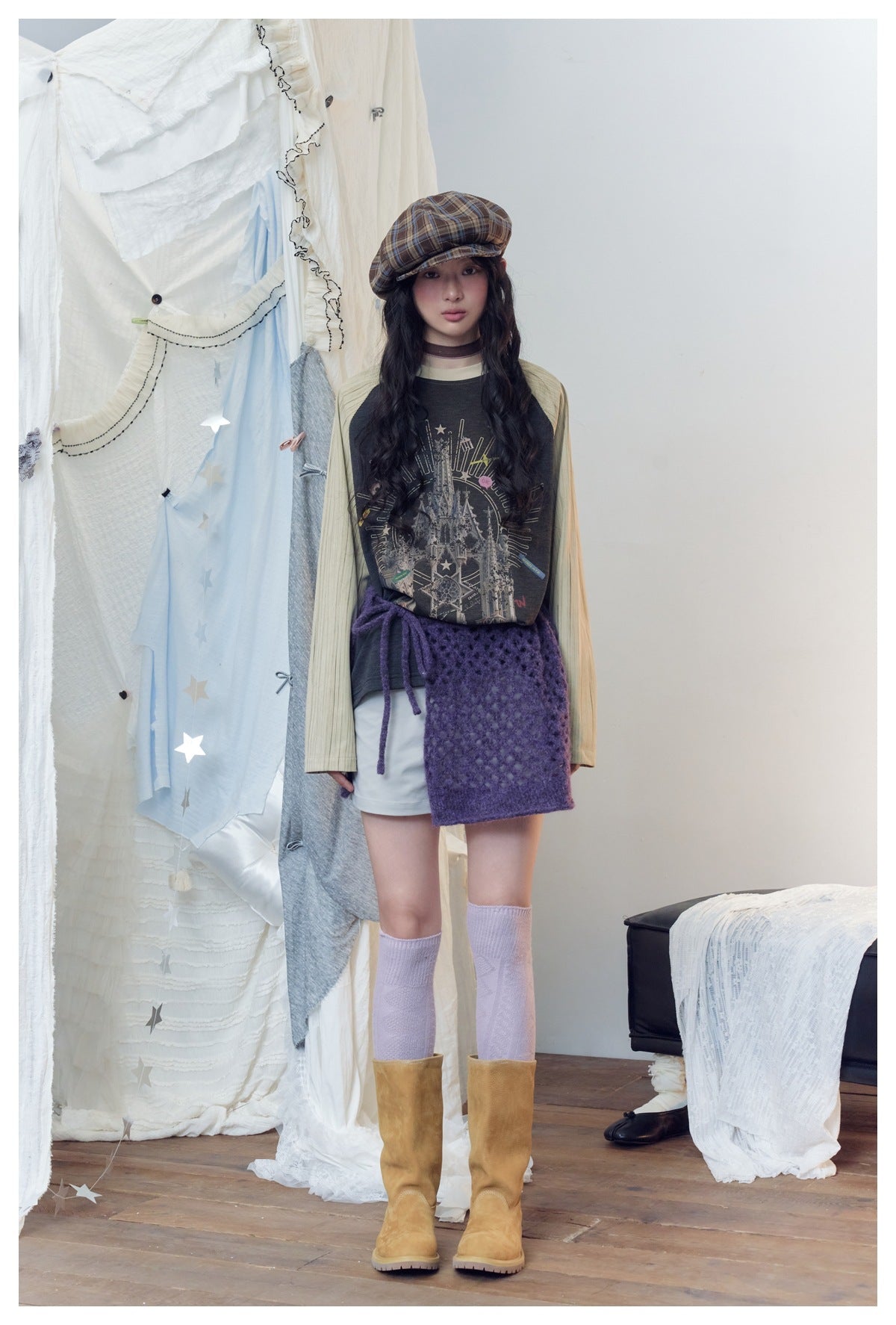 Model wearing the castle-print raglan sweatshirt with a plaid cap, purple knit skirt, lilac socks and tan boots