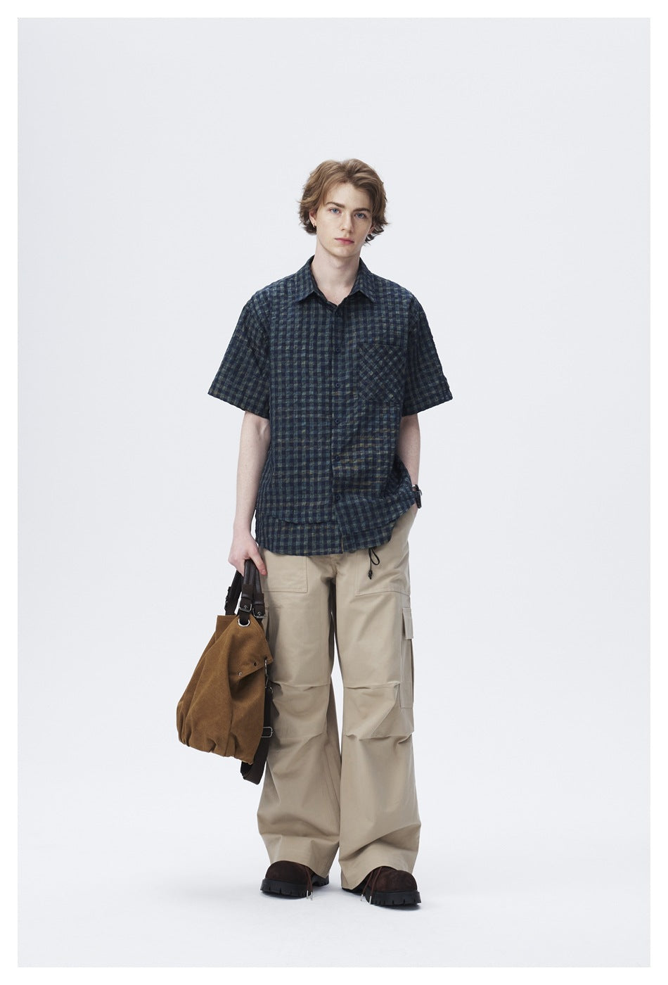 Model wearing dark check short-sleeve shirt with beige cargo pants and holding a brown suede bag, full-length view