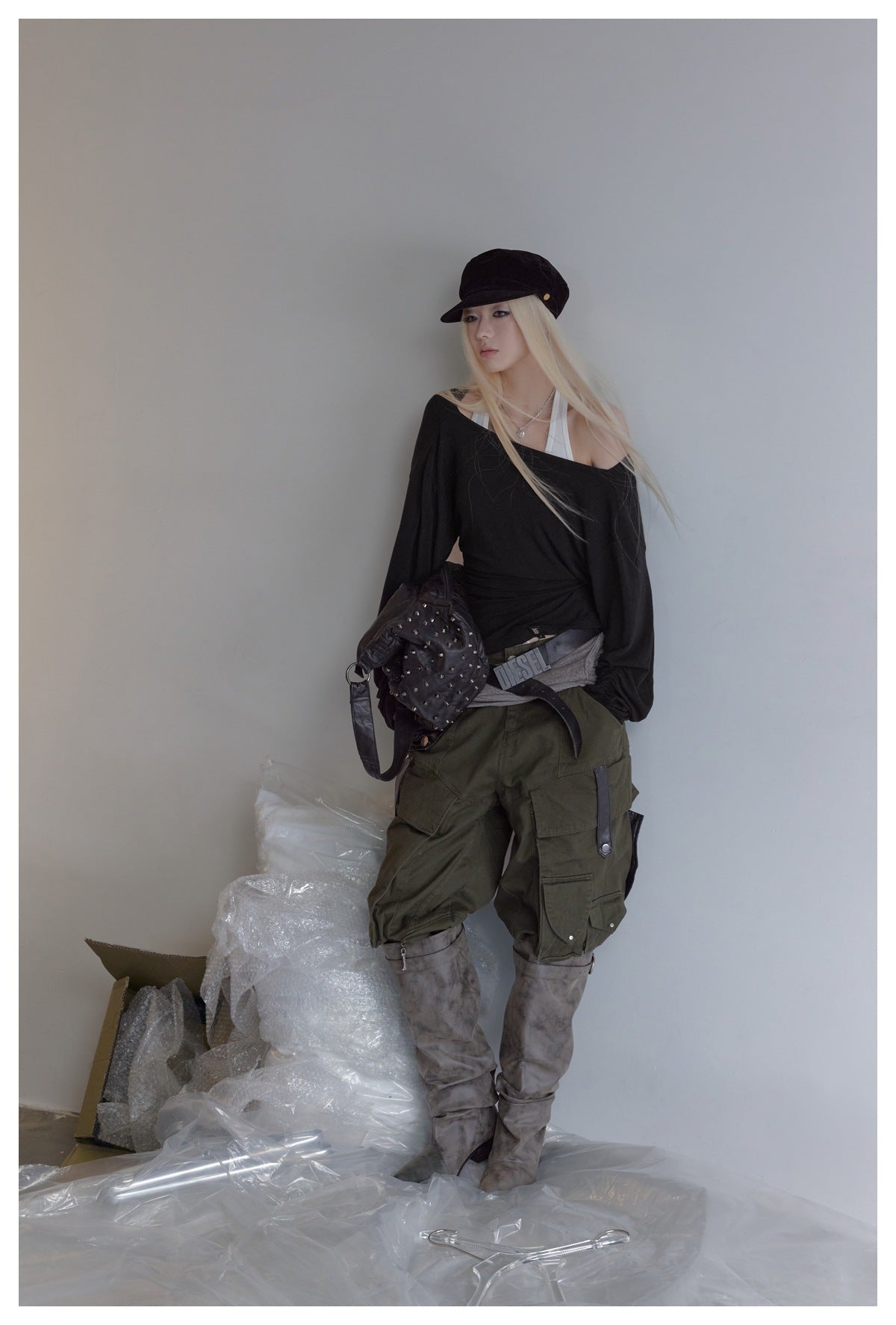 Model leaning on a wall in the black draped top with white halter layer, cap, studded bag, cargo pants and gray boots