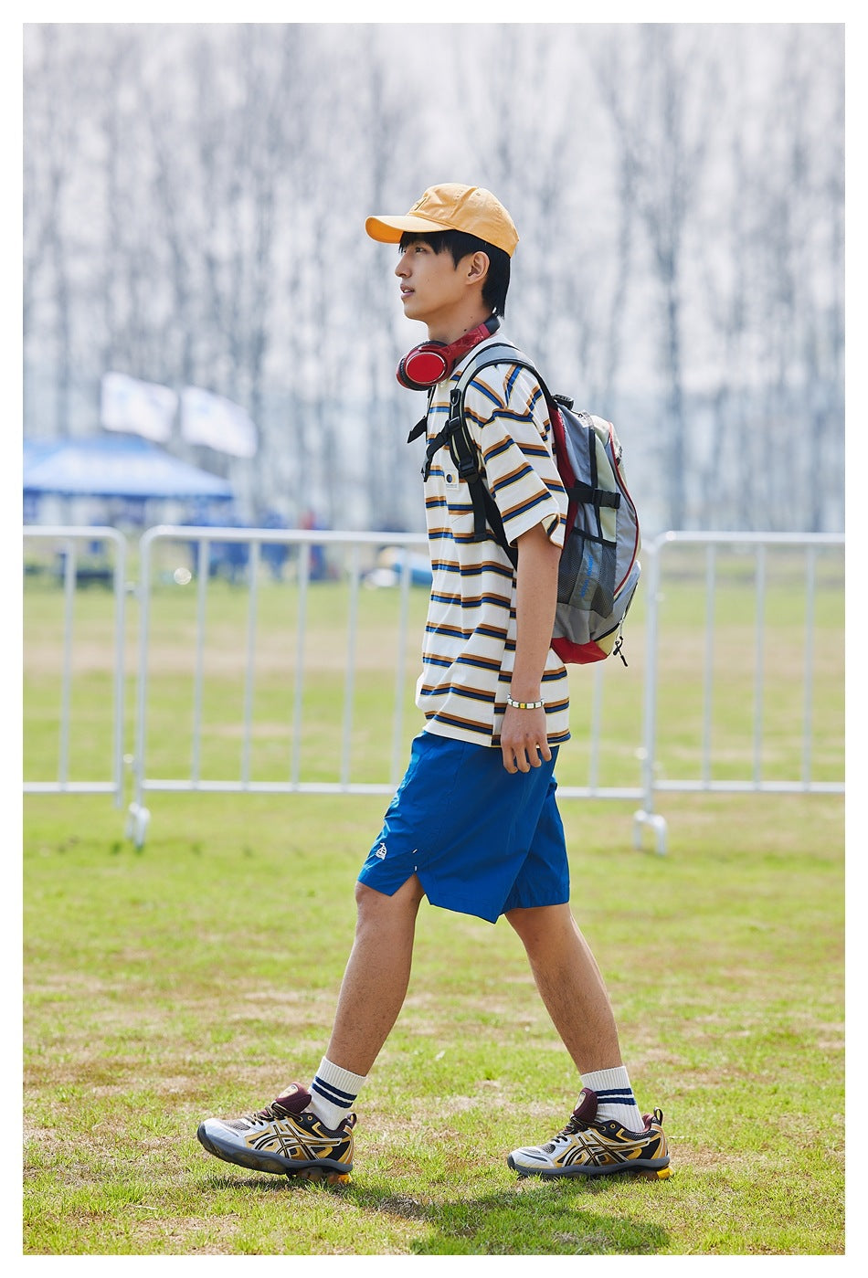 Side view of a model walking in the white striped pocket T-shirt with blue shorts, cap and backpack on a grass field
