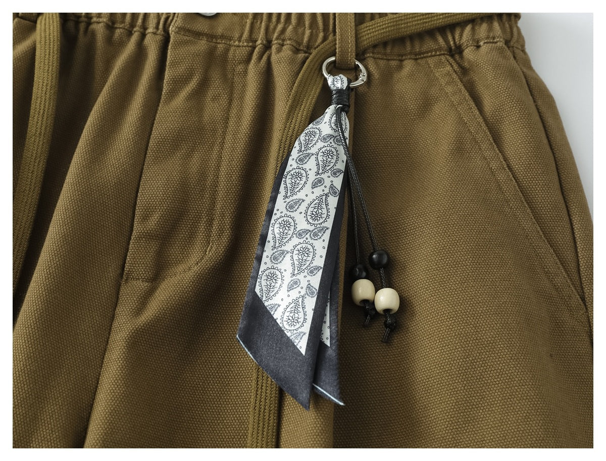 Close-up of olive-brown trousers showing the gathered waistband, front pocket and paisley-patterned hanging accessory with