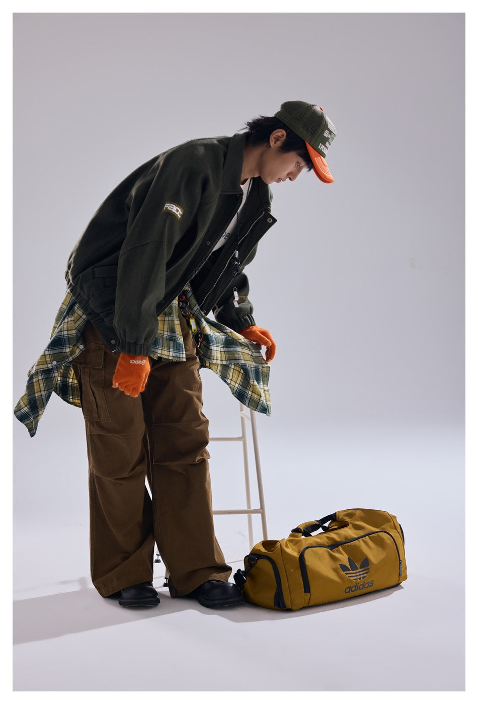 Model in a dark olive jacket and brown trousers bends beside a mustard-yellow duffel bag.