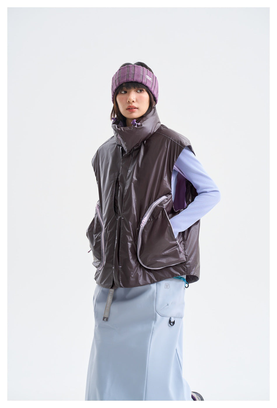 Model wearing a charcoal-gray sleeveless vest with large zip pockets over a lavender top and pale blue skirt.