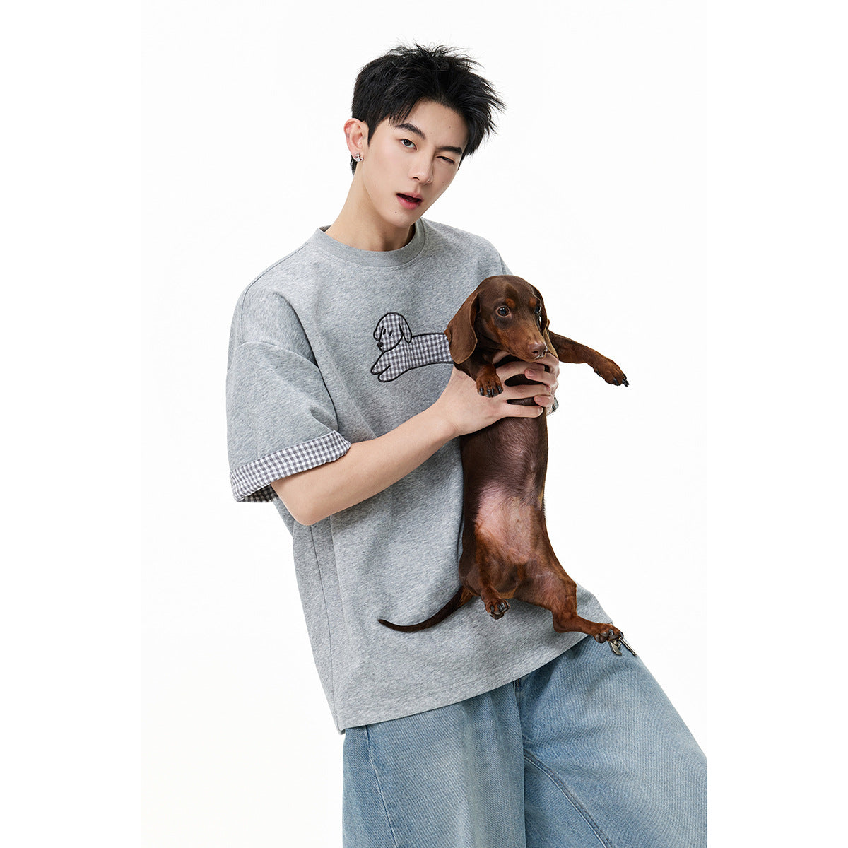 Man wearing a light gray T-shirt with a dog graphic and checked sleeve cuffs, holding a brown dachshund.