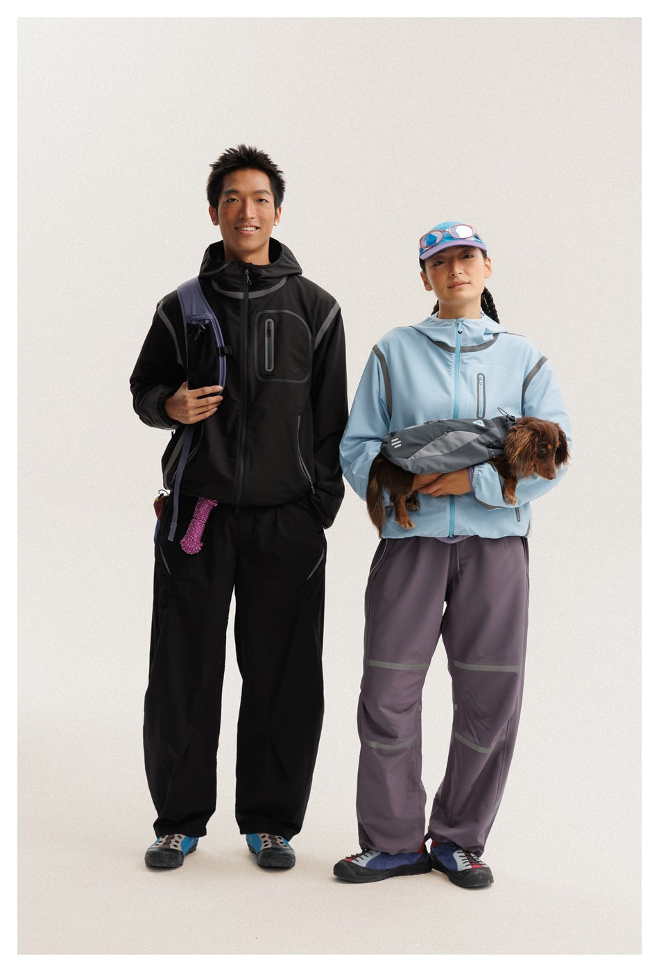 Two models wear hooded jackets in black and light blue, paired with black and muted purple trousers.
