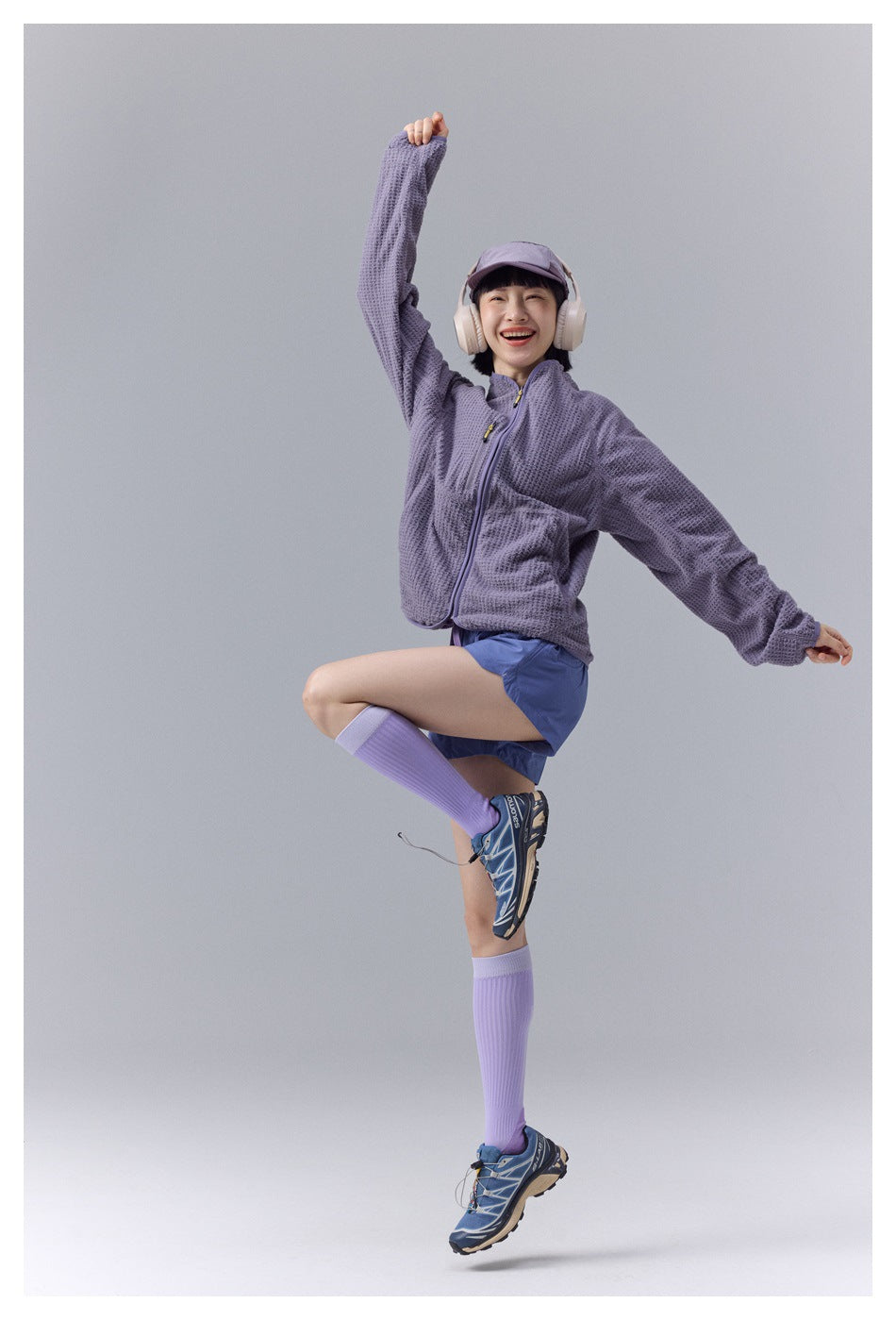 Model in a lavender zip jacket and blue shorts, posing energetically in a full-body front view.