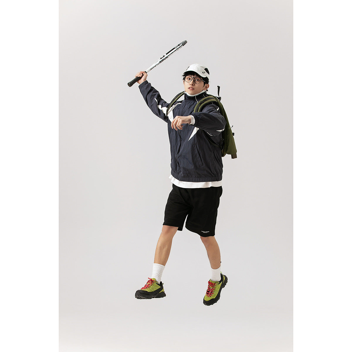 Model in a black-and-white paneled jacket and black shorts poses with a tennis racket raised overhead.