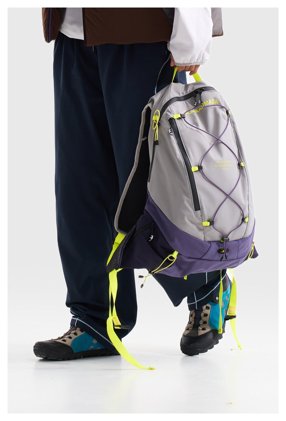 A person holds a gray and purple backpack with neon yellow accents, front bungee cord, and zip pockets.