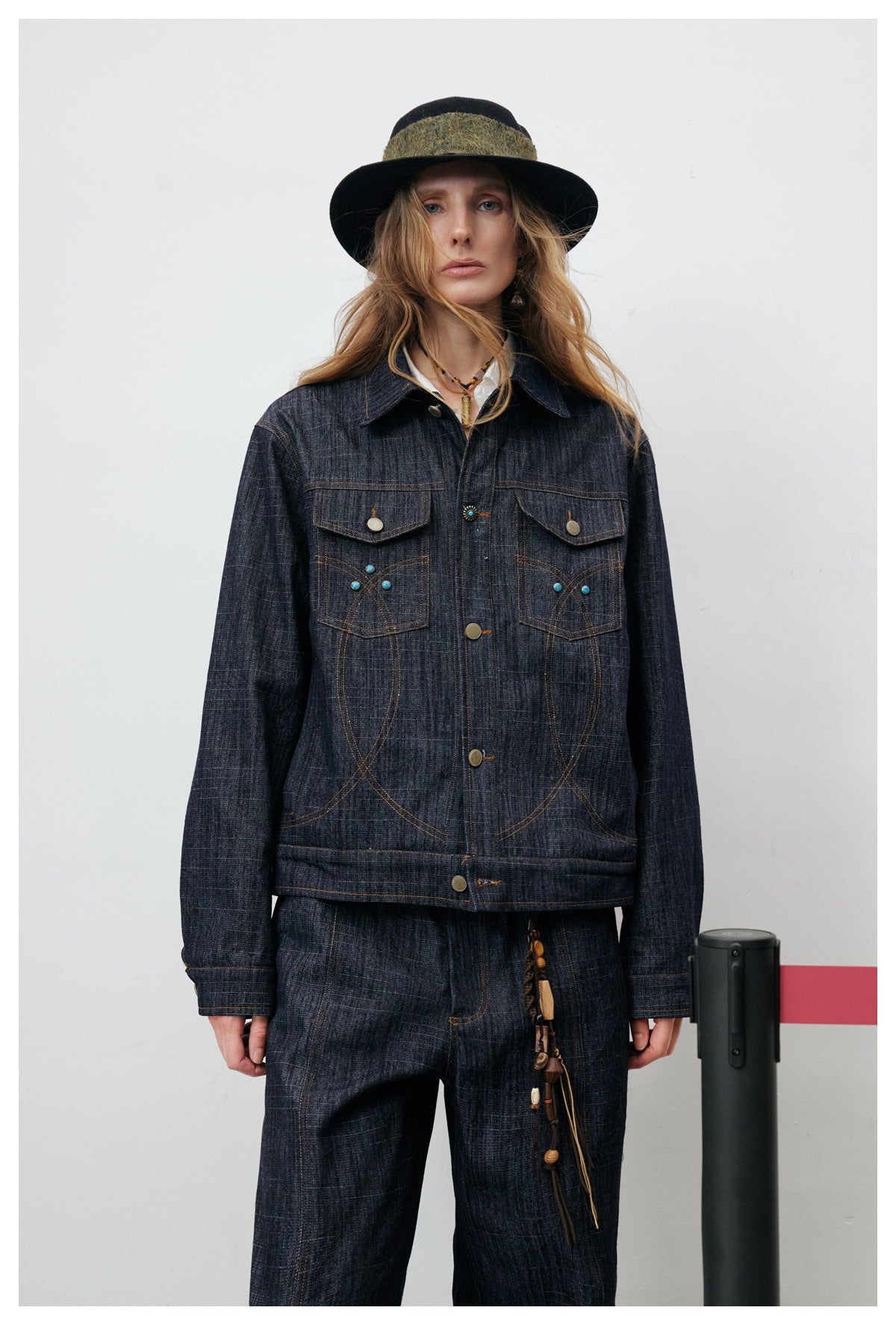 Model wearing the front of a dark indigo denim jacket with flap pockets, turquoise accents, and matching trousers.