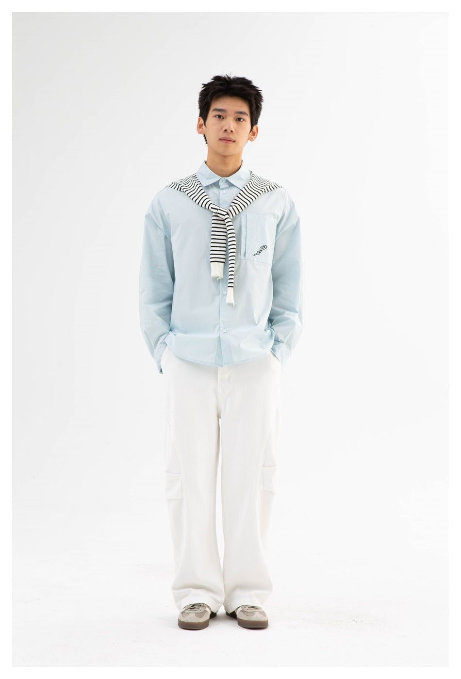 Model wearing a light blue button-front shirt with chest pocket, striped sweater over shoulders, white pants and sneakers