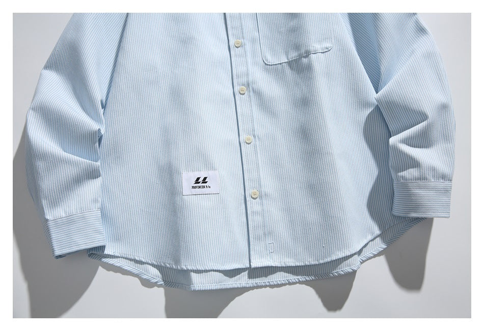 Light blue and white striped button-up shirt hanging against a pale wall, with a chest pocket and small sewn logo label.