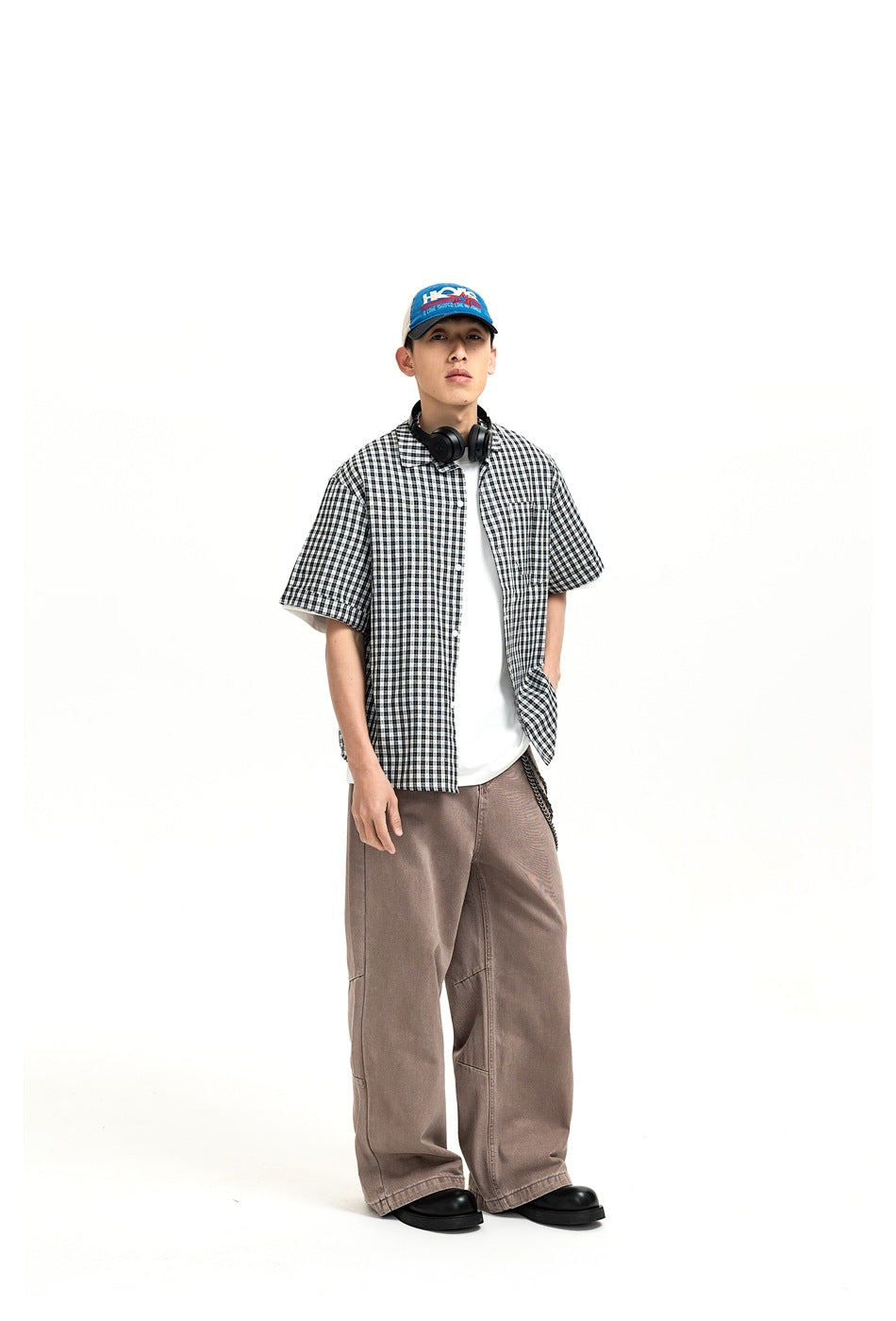 Man wearing brown wide-leg jeans with a checked short-sleeve shirt, white tee, cap and chain, standing front view