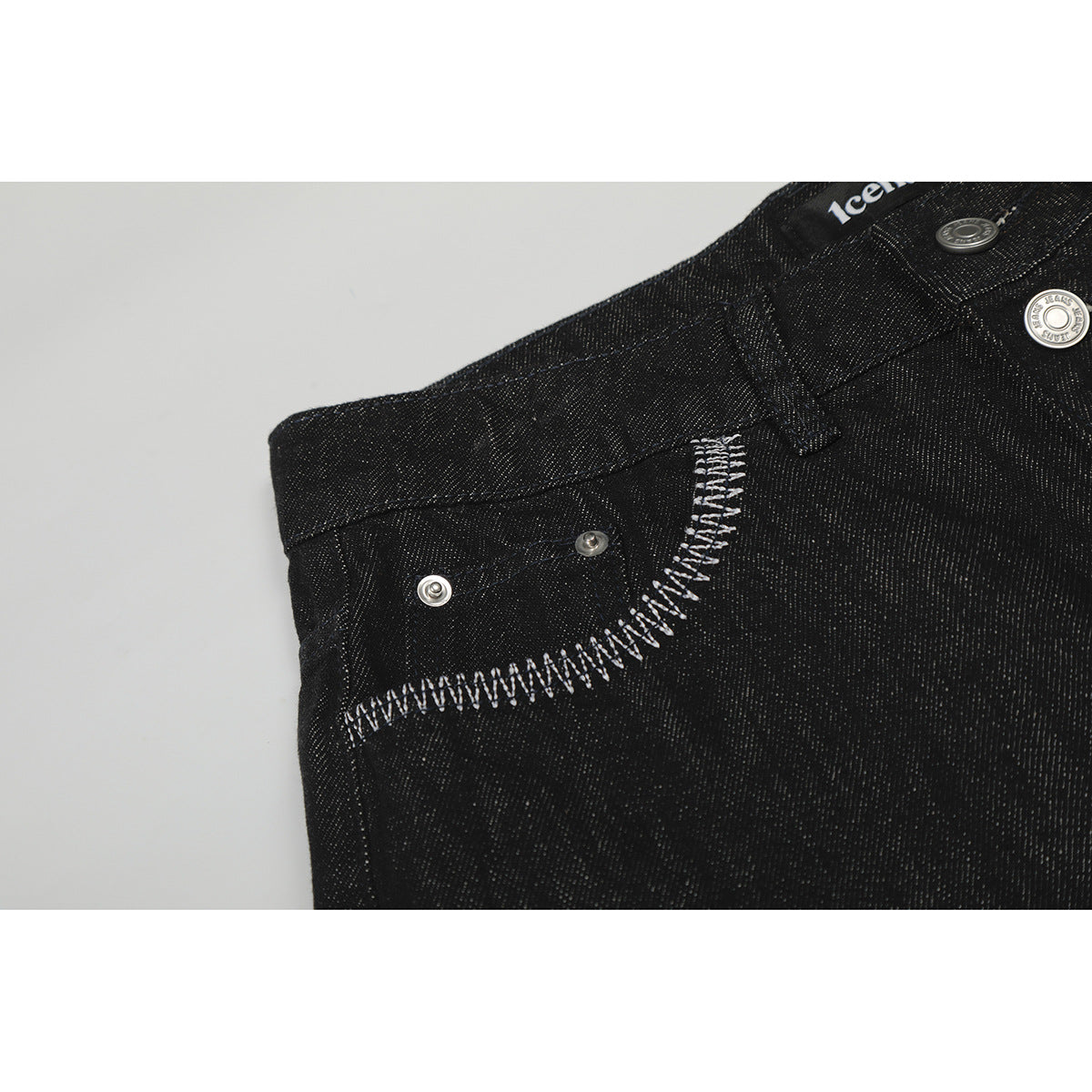 Close-up of black jeans showing a curved front pocket, metal rivets, and bold white stitching.