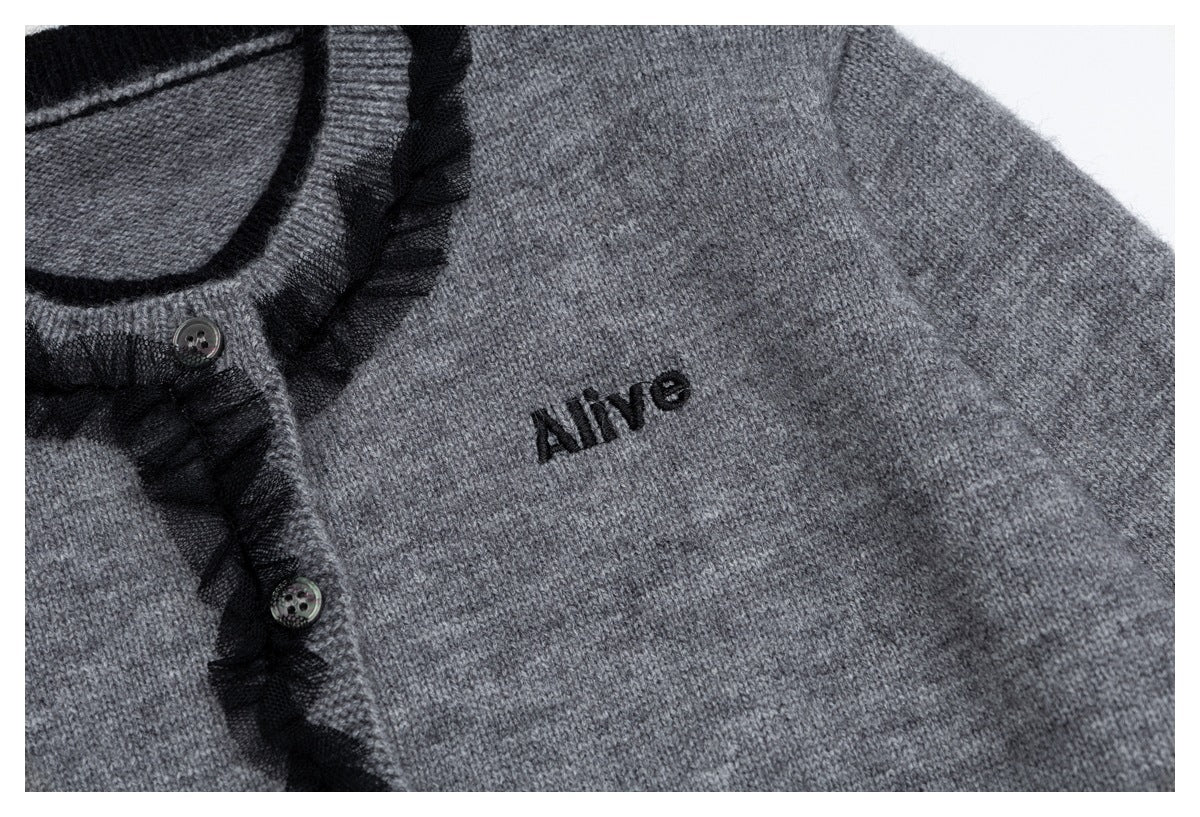 Close-up of a gray knit cardigan with black ruffle trim, buttons, and small black embroidered “Alive” lettering.