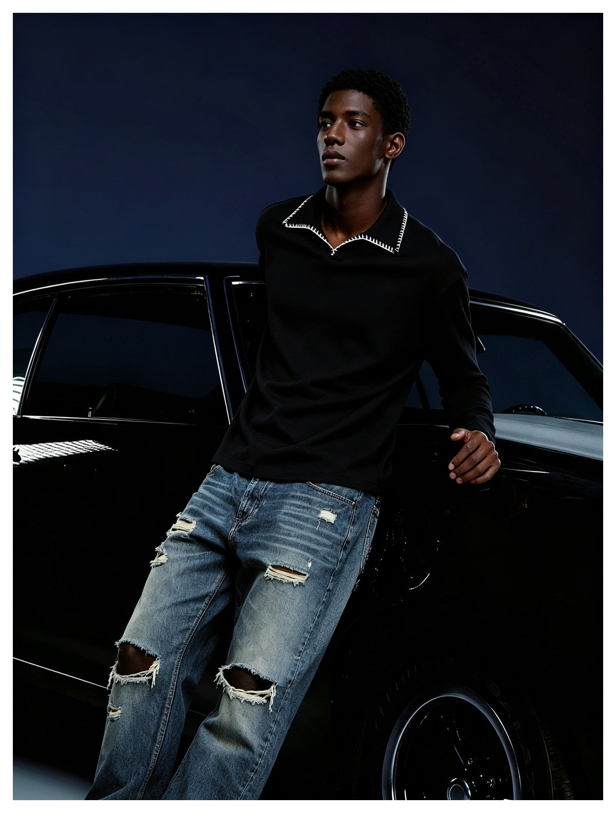 Man in a black stitched-collar long-sleeve top and ripped jeans leaning on a black car against a dark blue backdrop