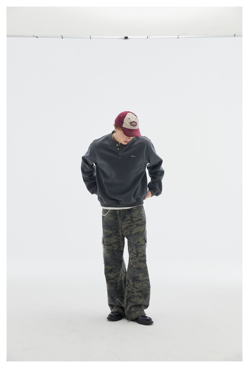 Model in a charcoal three-button henley sweatshirt and loose camouflage cargo pants, adjusting the sweatshirt hem.