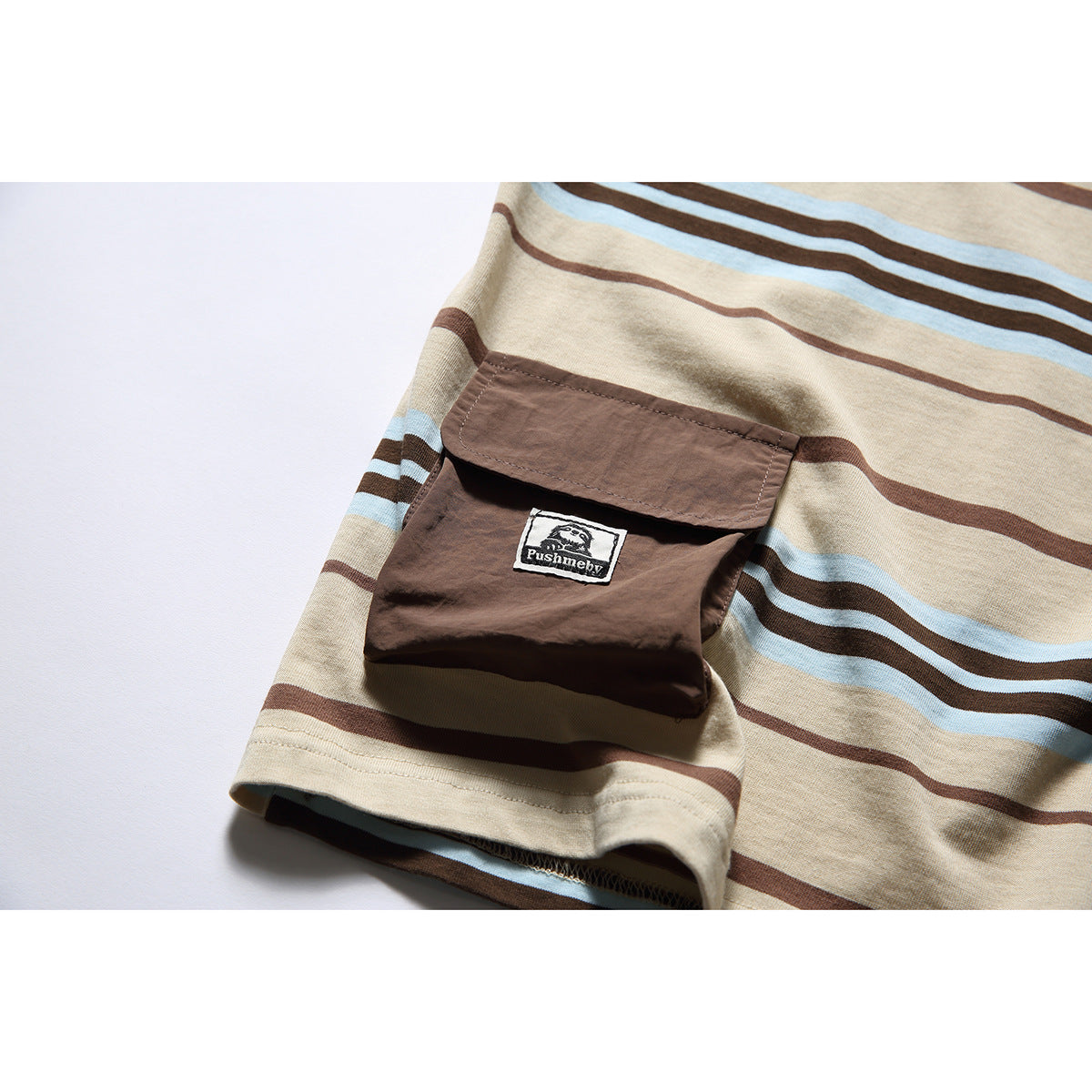 Close-up of a brown flap pocket with a small sewn label on a beige T-shirt with brown and light blue stripes.