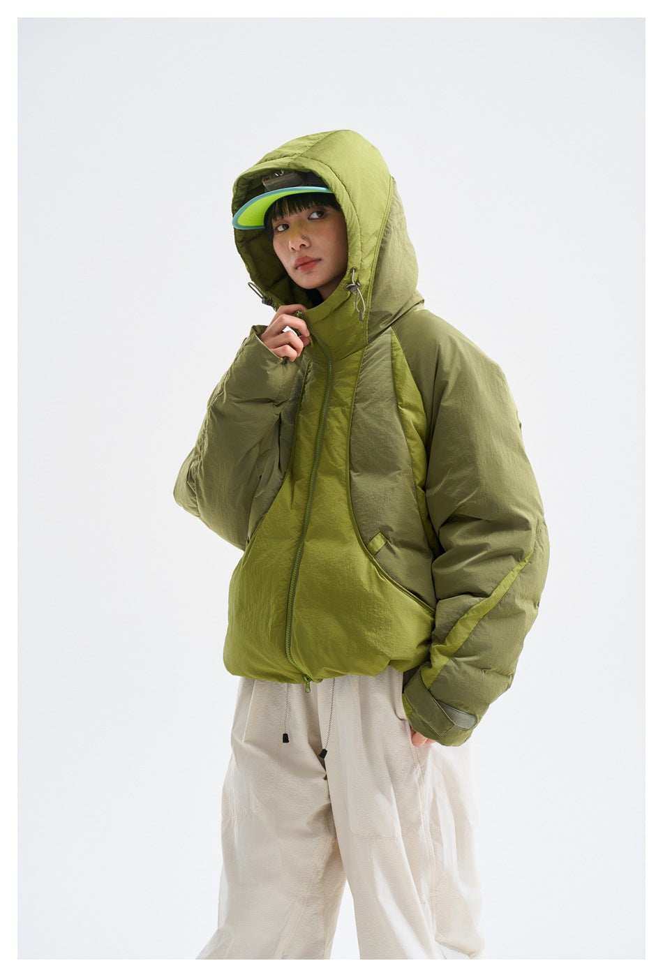Closer view of a model in an olive and lime-green hooded jacket with the hood raised and a cap underneath.